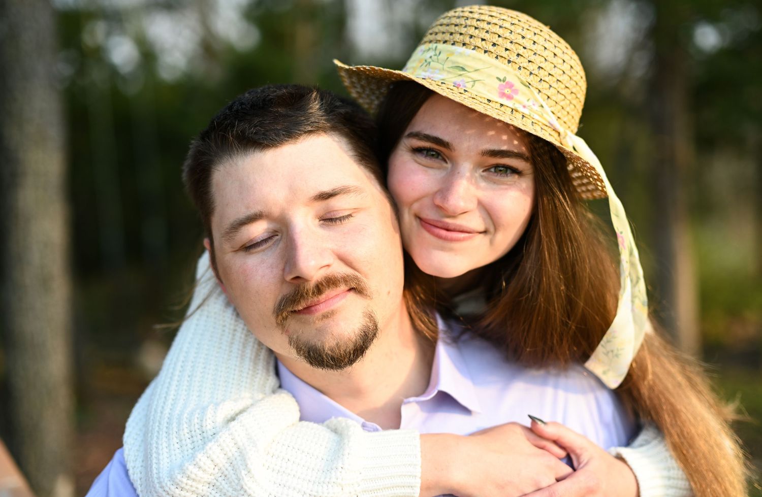 A smiling young woman in a straw hat hugs a bearded man from behind in a warm outdoor portrait.
