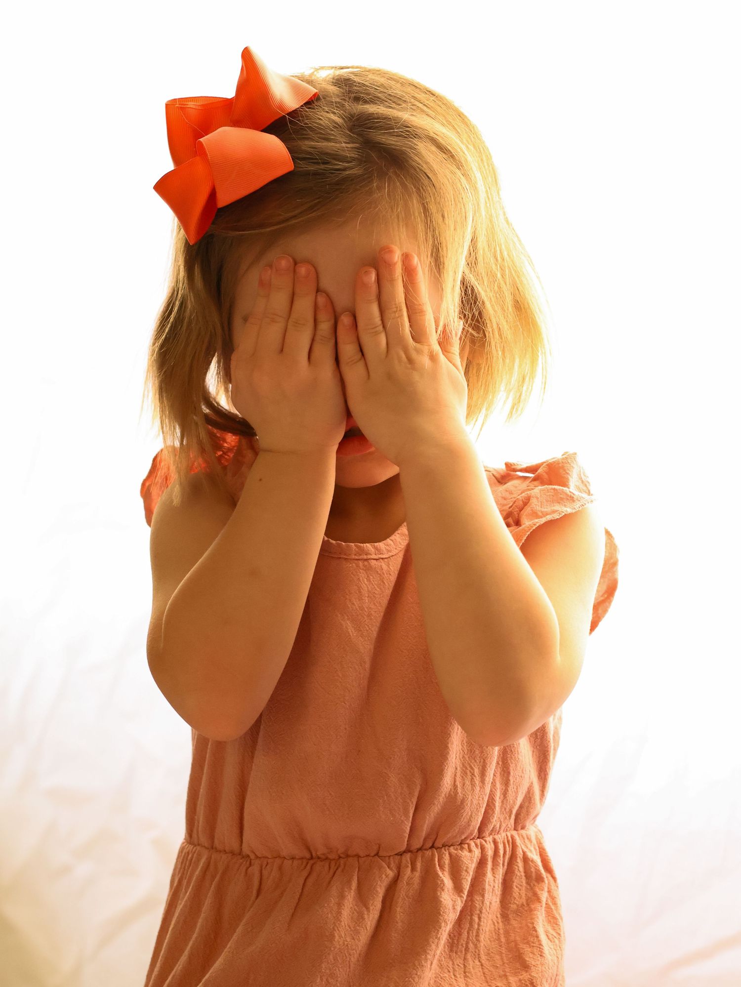 Young girl in orange dress covering her face with both hands playfully.