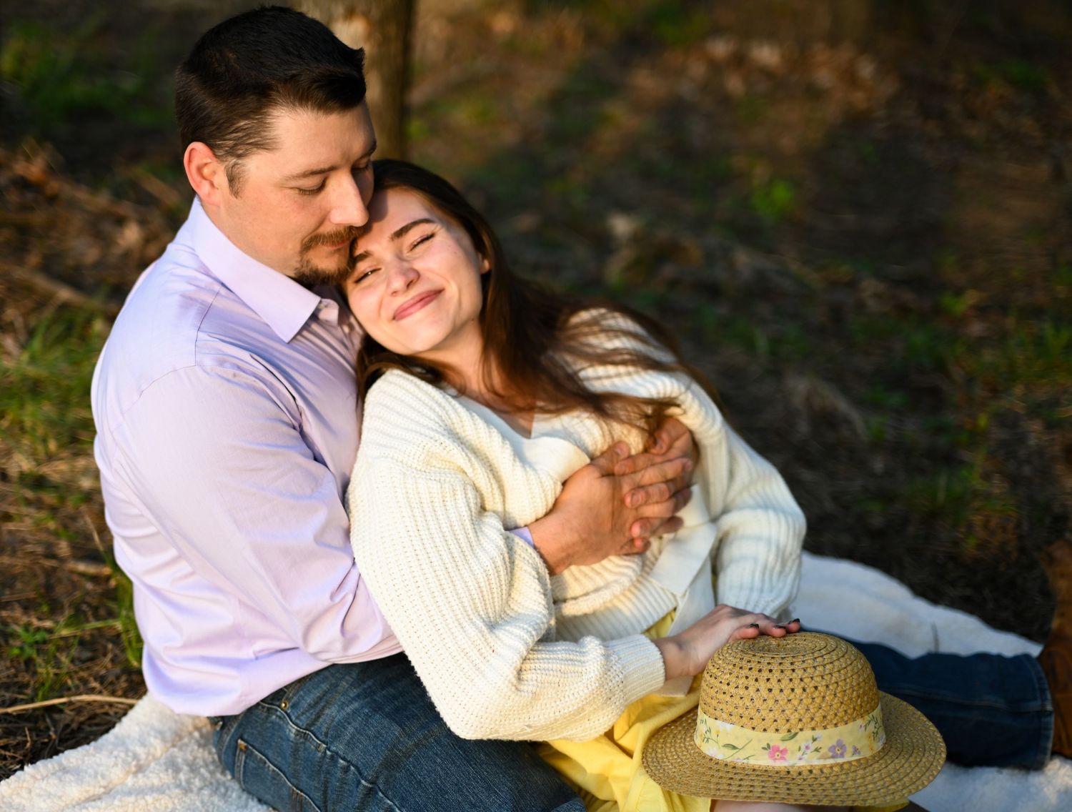 A couple sits closely on a blanket outdoors, eyes closed, embracing peacefully with a straw hat nearby.
