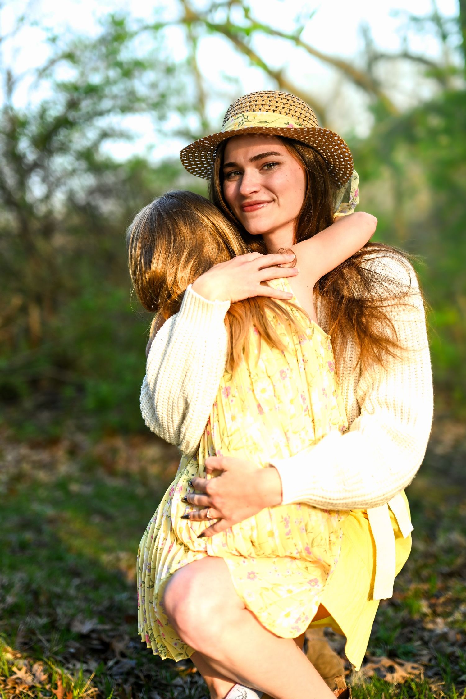 A smiling woman in a straw hat hugs a young girl in a yellow dress outdoors in golden sunlight.