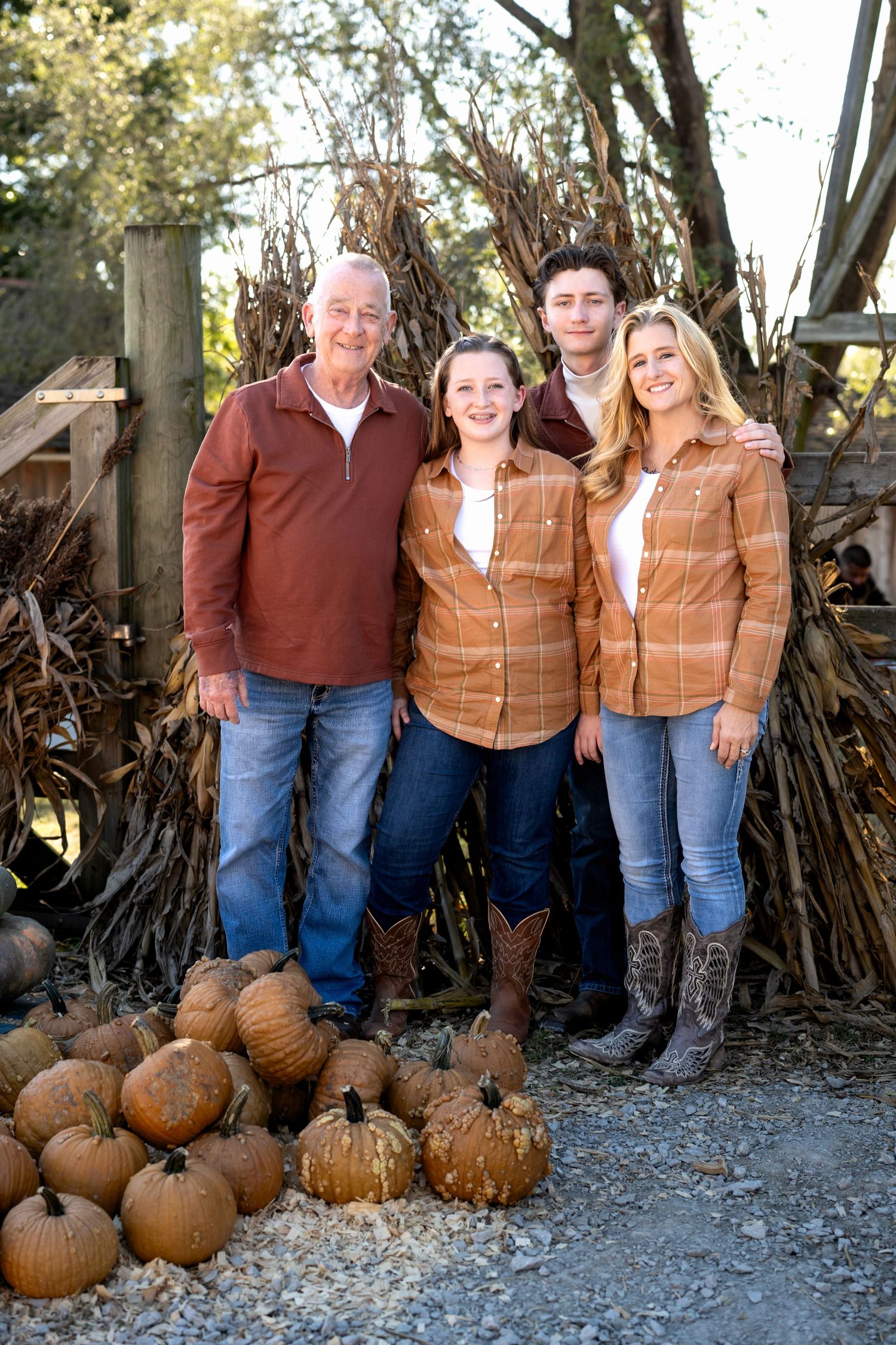Family of four in matching rust and plaid flannel shirts pose beside corn stalks and a pile of pumpkins for an autumn portrait.
