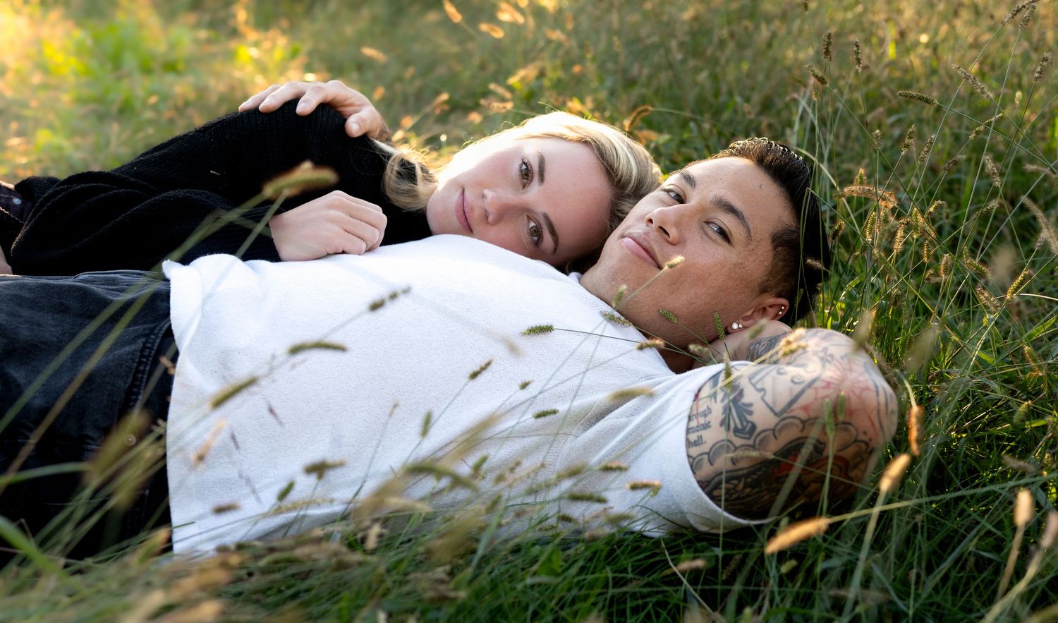 Smiling couple lying close together in a sunlit grassy field, the man in a white shirt with an arm tattoo and the woman resting beside him.
