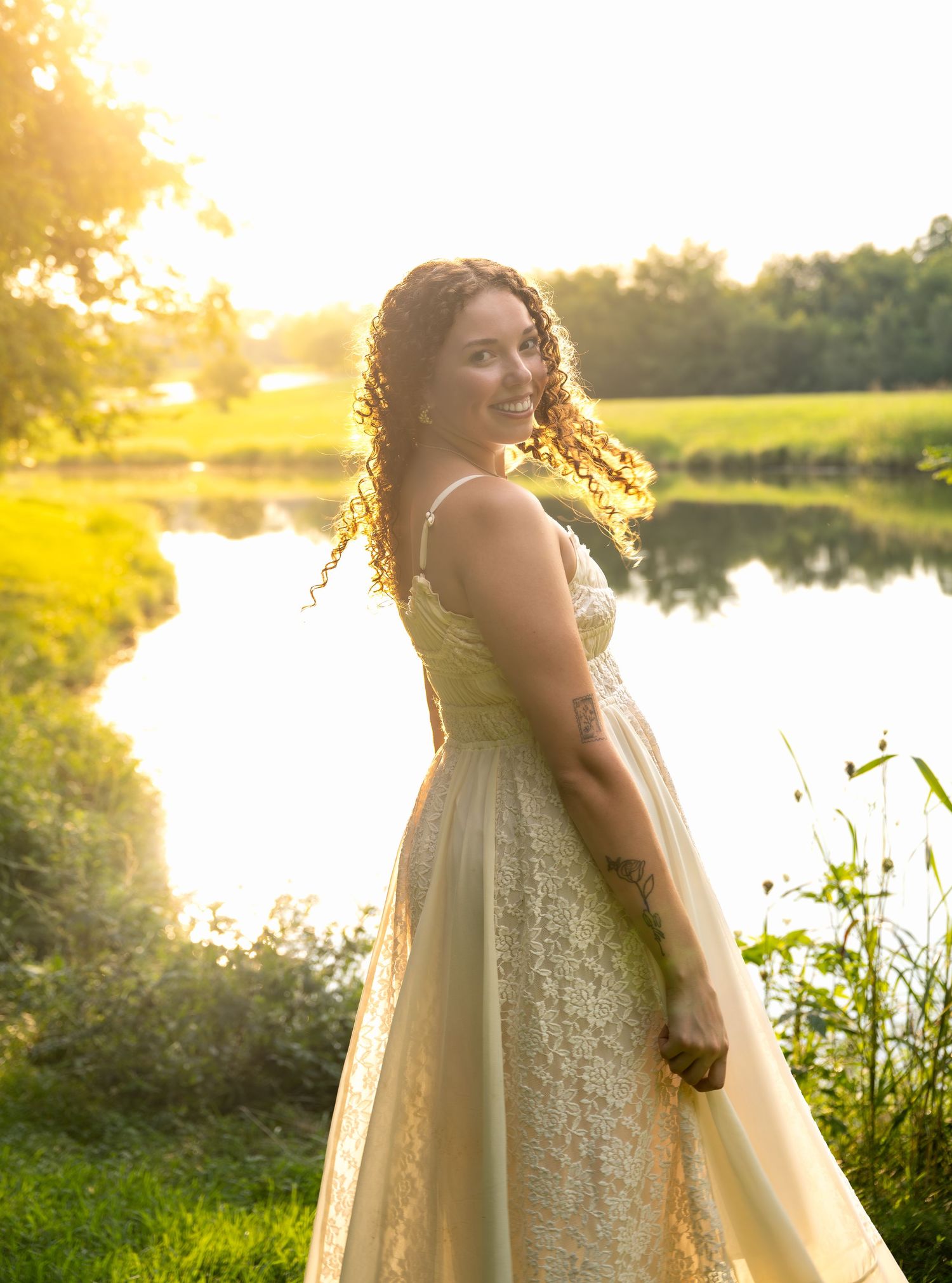 lifestyle portrait of a Young woman in golden lace gown twirling by a sunlit pond at sunset with curly hair flowing.
