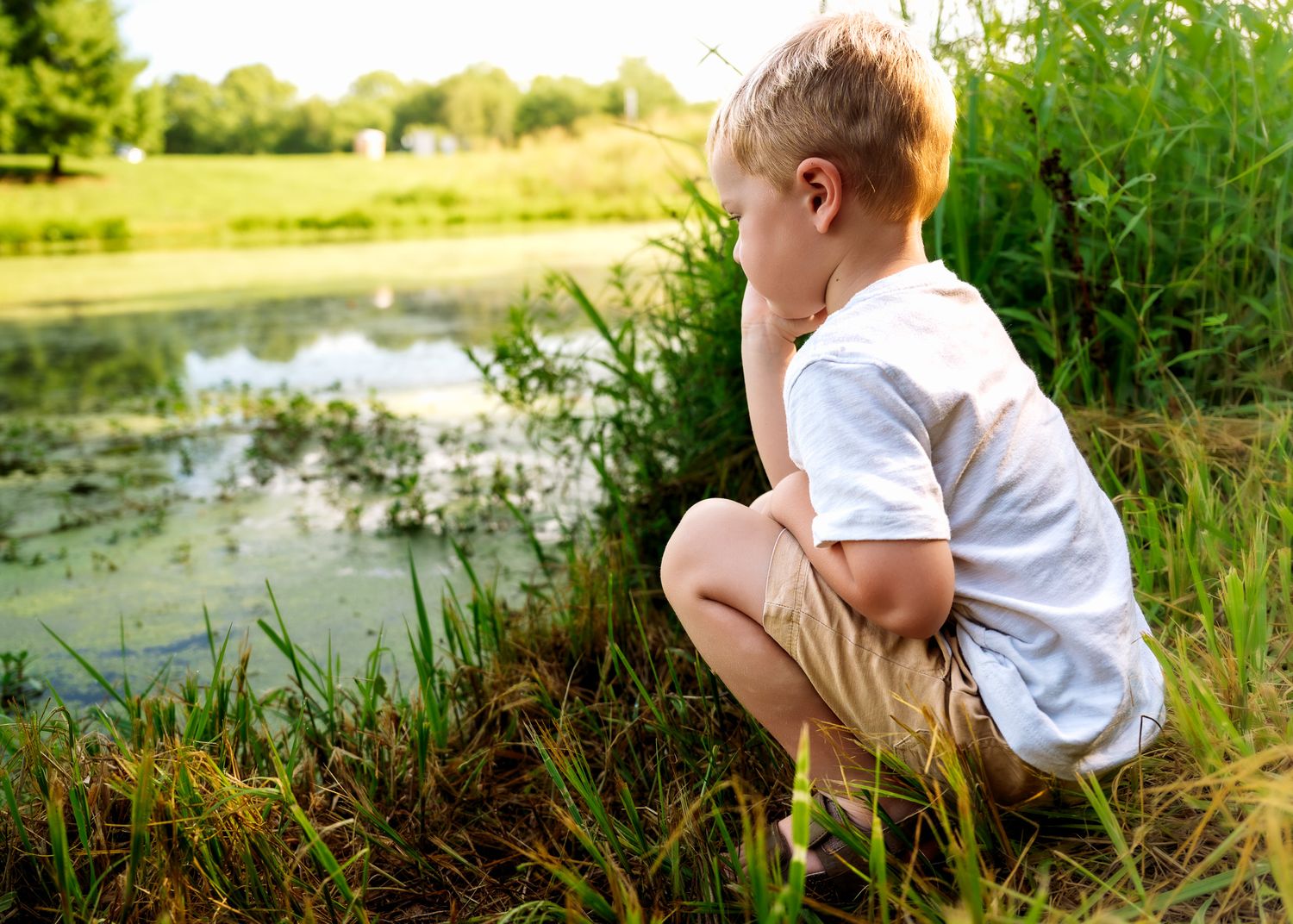 Young blonde boy crouching by a peaceful pond, exploring nature in a sunny green park setting. faceless family photography in Leavenworth KS