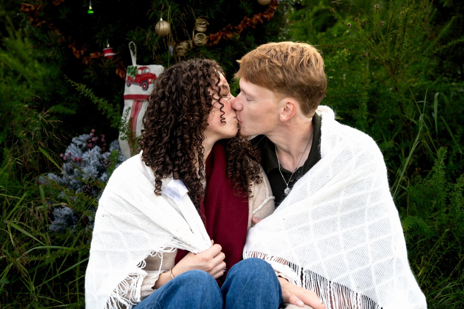 A young couple wrapped in a white blanket shares a kiss outdoors near festive Christmas decorations and greenery.