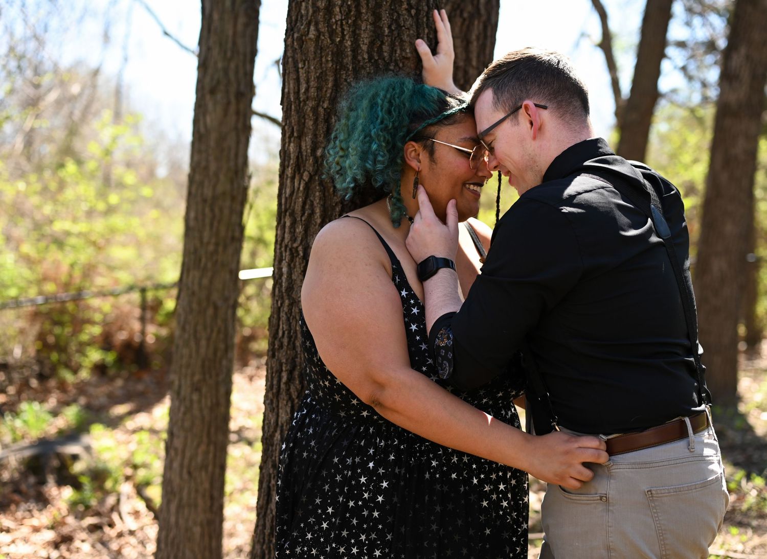 Interracial couple leaning in close together against a tree in a wooded outdoor setting.