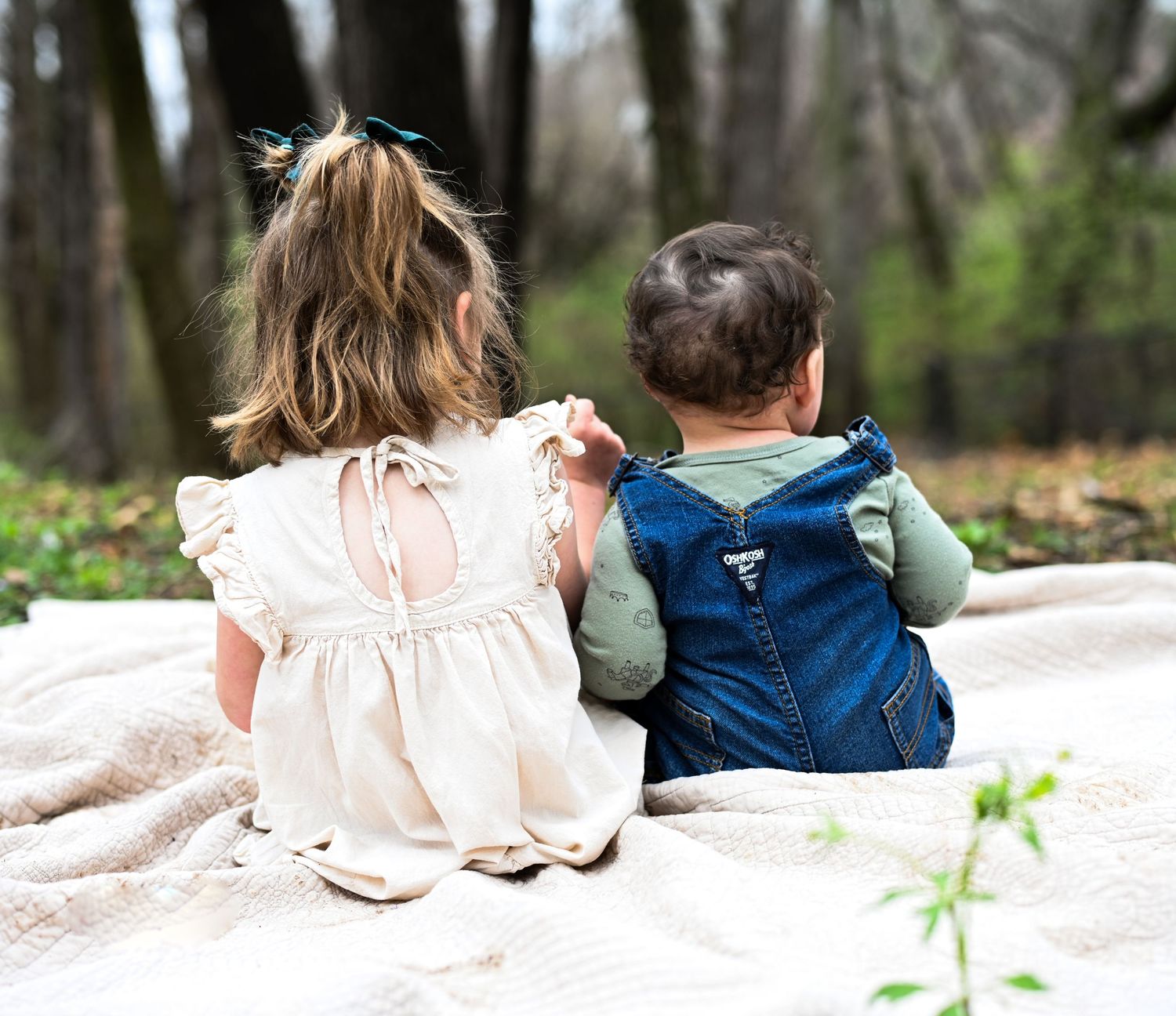 Two young children in white dress and denim overalls sitting together on blanket in forest park.