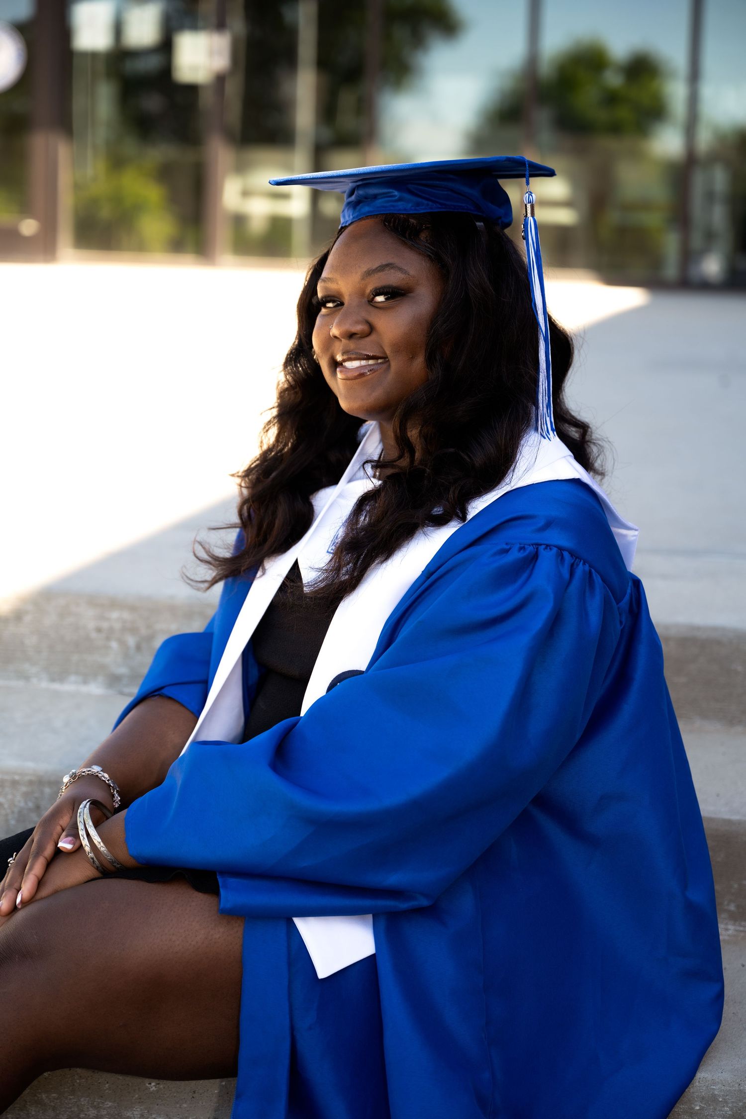 portrait of a Smiling graduate in blue cap and gown sitting on steps celebrating academic achievement outdoors.