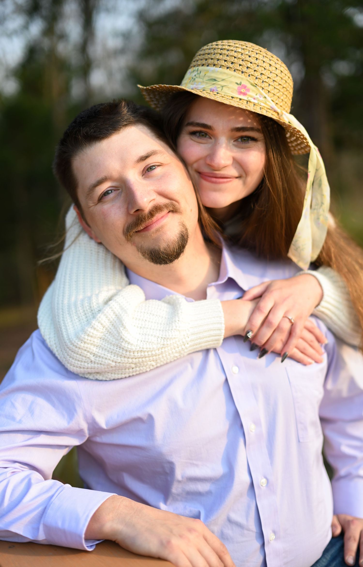 A happy couple poses together outdoors, woman in straw hat resting chin on mans cheek smiling.
