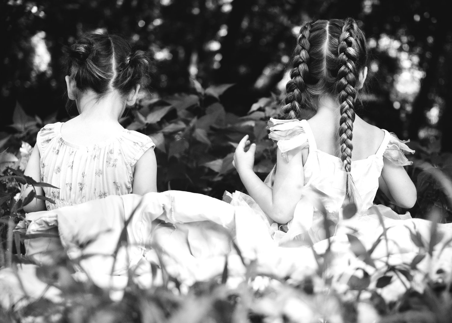 Two girls with braided hair in white dresses sit among lush green foliage in a black and white photo.