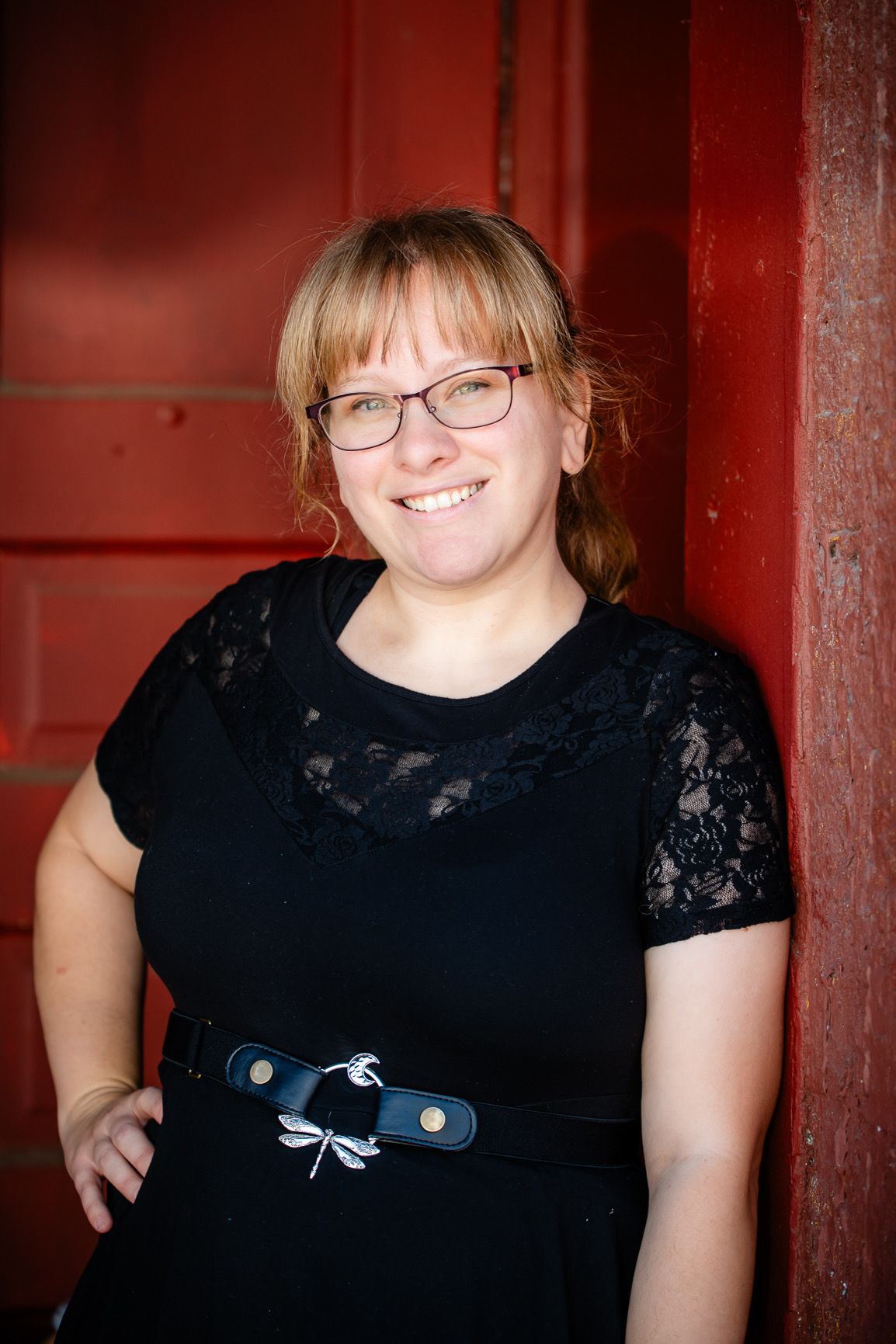 Smiling woman with glasses in black lace dress leaning against a rustic red barn door.