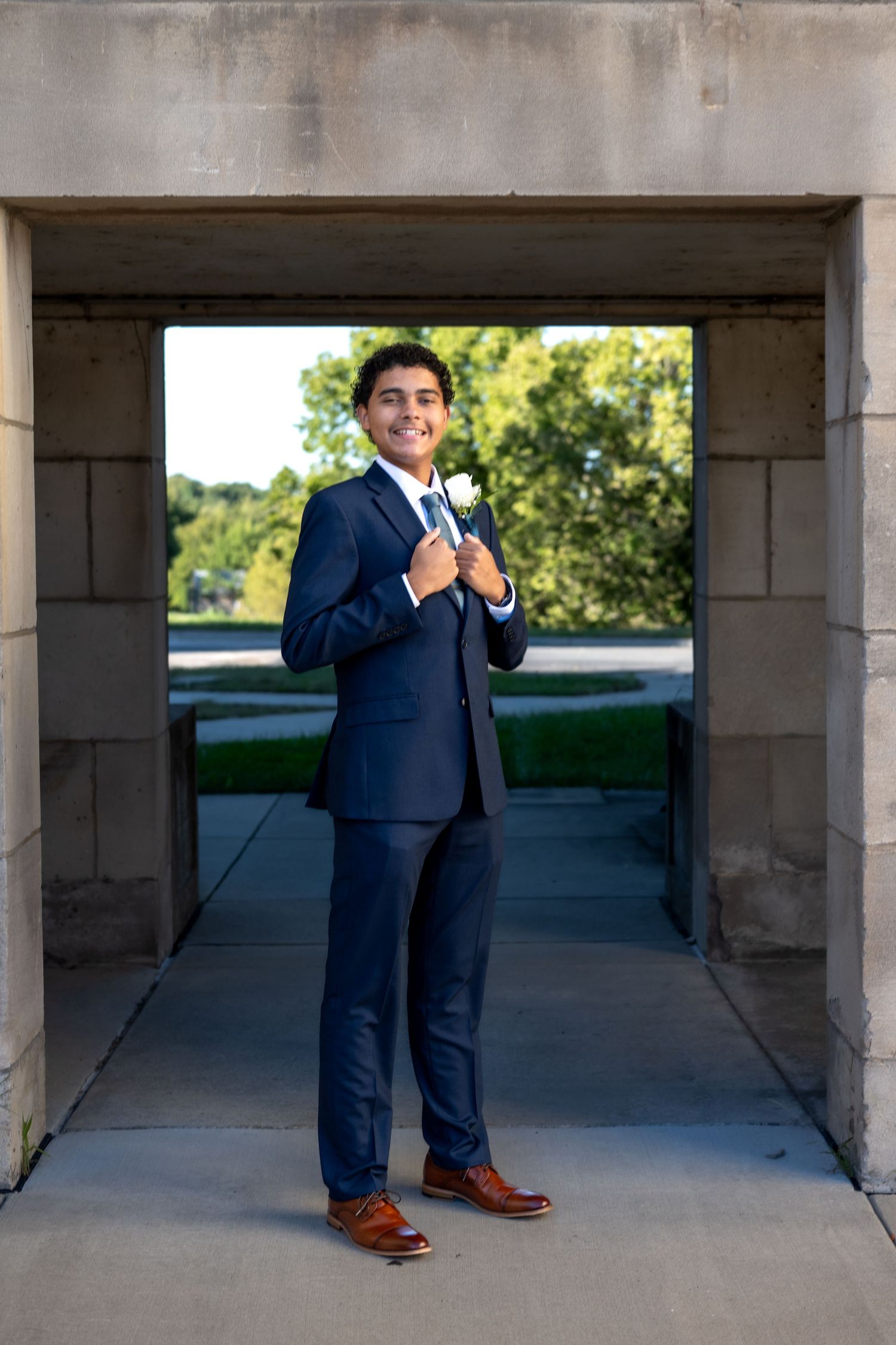 Young man in navy suit with white boutonniere poses confidently under stone archway outdoors.