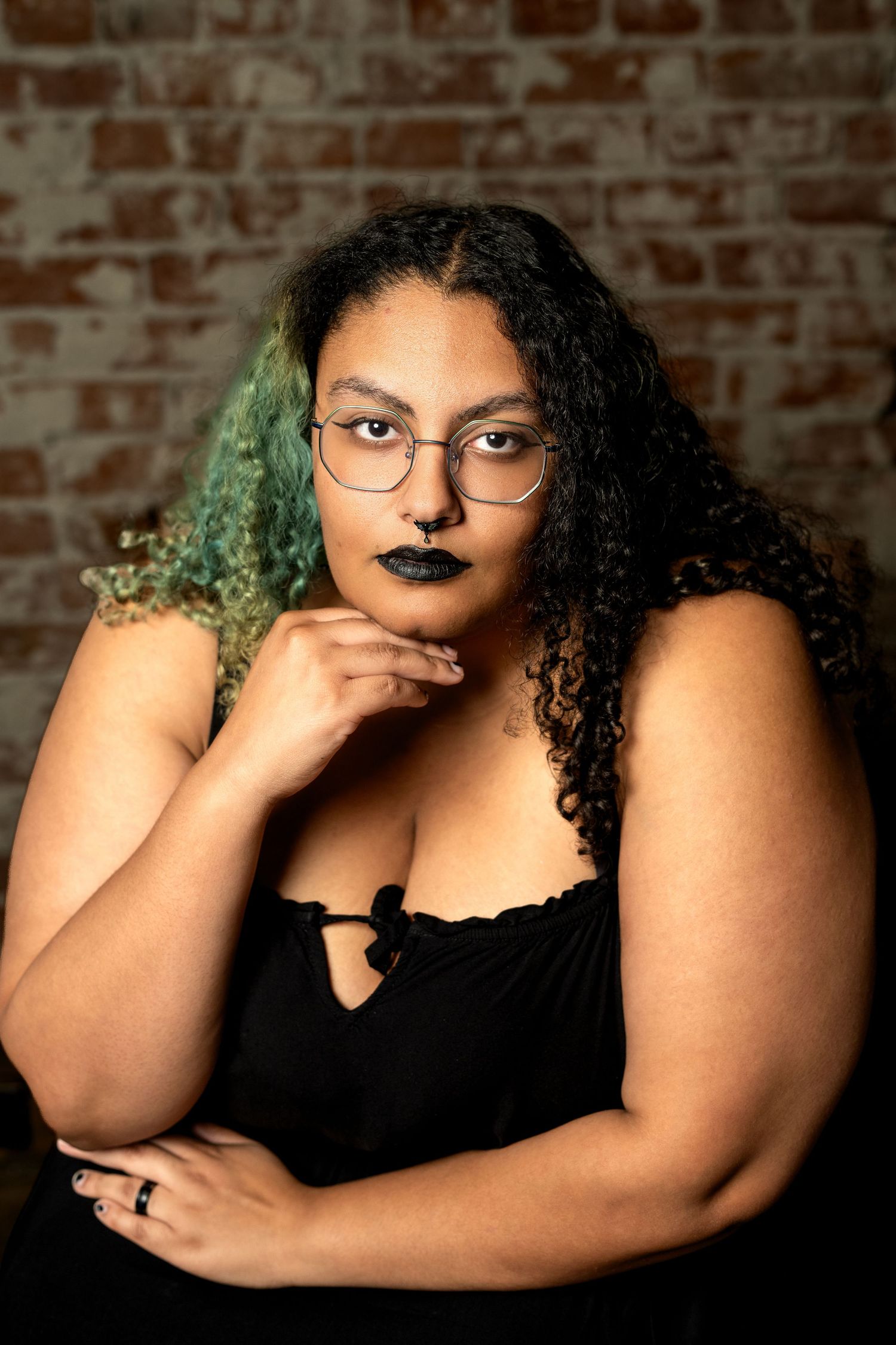 Plus-size woman with curly green-tipped hair, black lipstick, glasses, posing confidently in black top against brick wall.