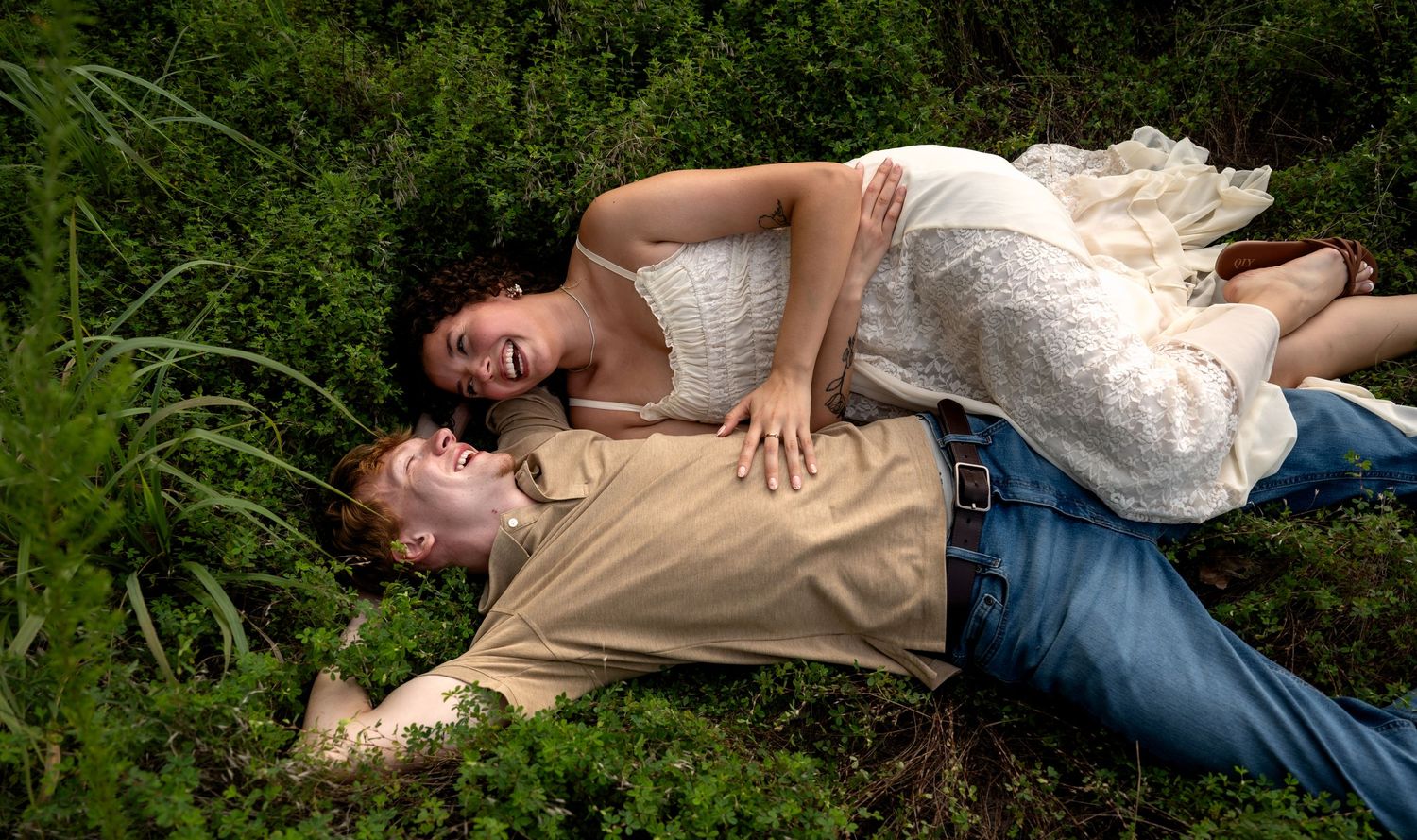A couple lies together in lush green grass, smiling and relaxed, with the woman in a white dress resting on the man.