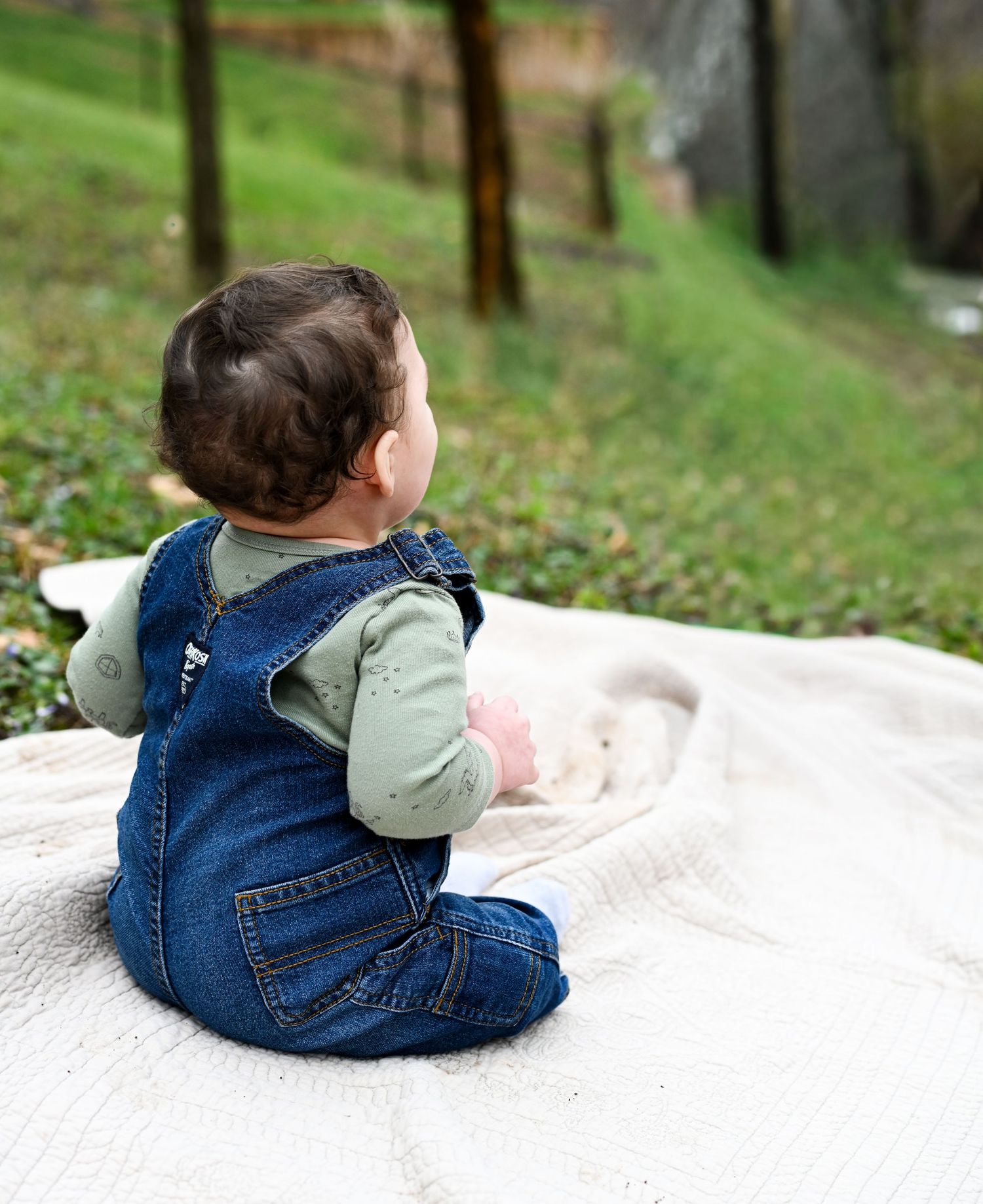Baby in denim overalls and sage green shirt sitting on white blanket outdoors in green park.