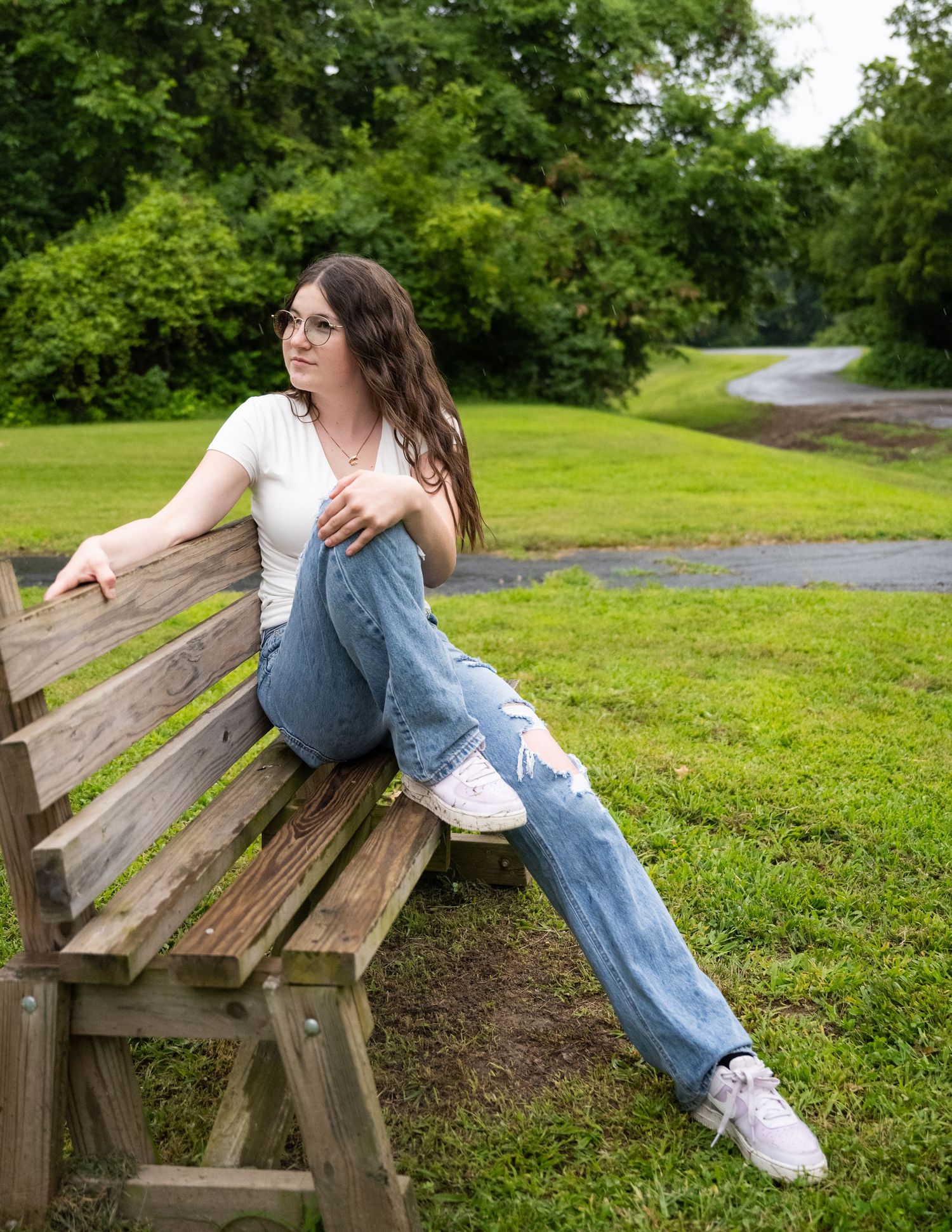portrait of a Teen girl in white top and ripped jeans sitting on park bench with winding path behind her.