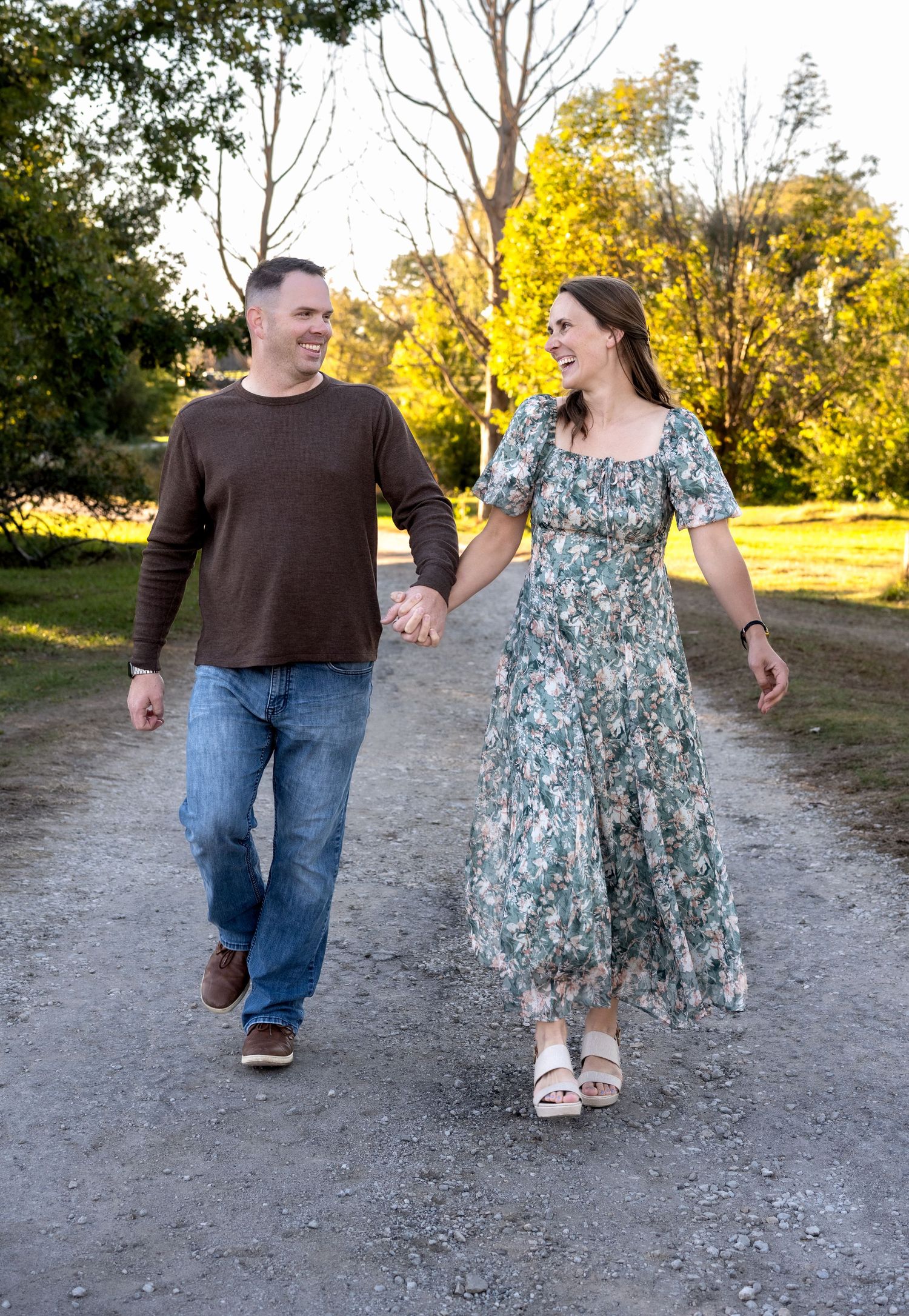A smiling couple holds hands and walks down a gravel path lined with autumn trees, he in a brown sweater and jeans, she in a floral dress.
