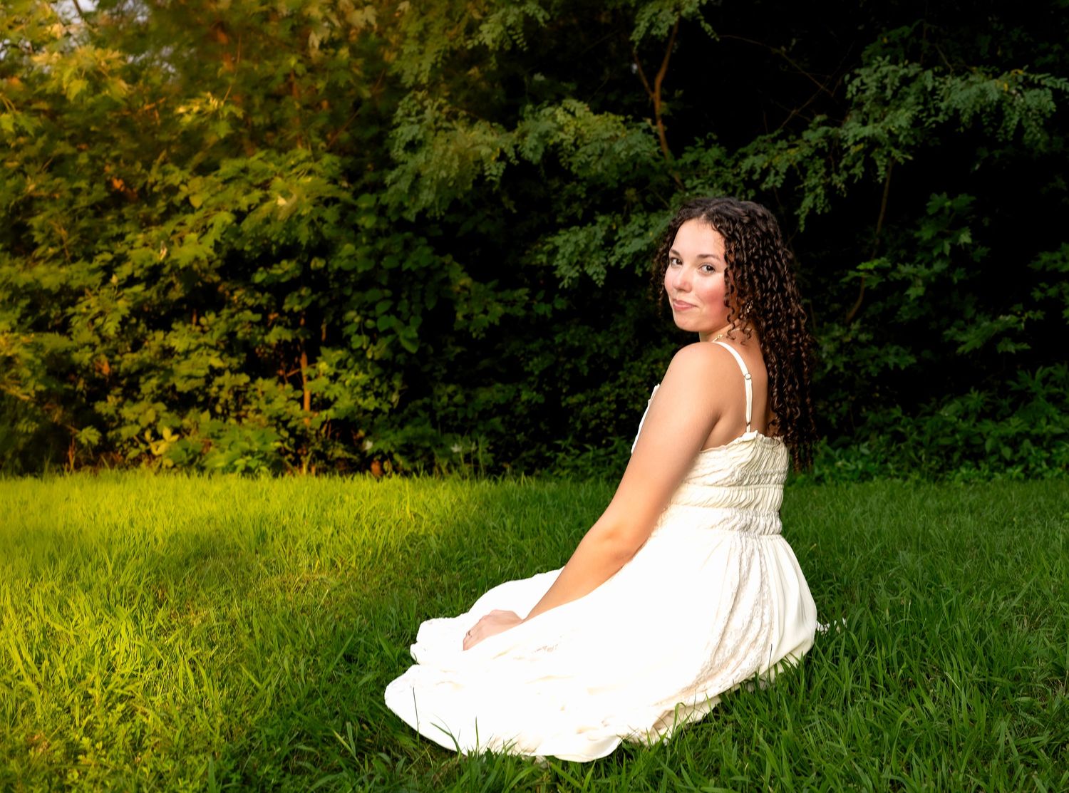 lifestyle portrait of a Young woman in white dress sitting on lush green grass, smiling over shoulder with trees in background.