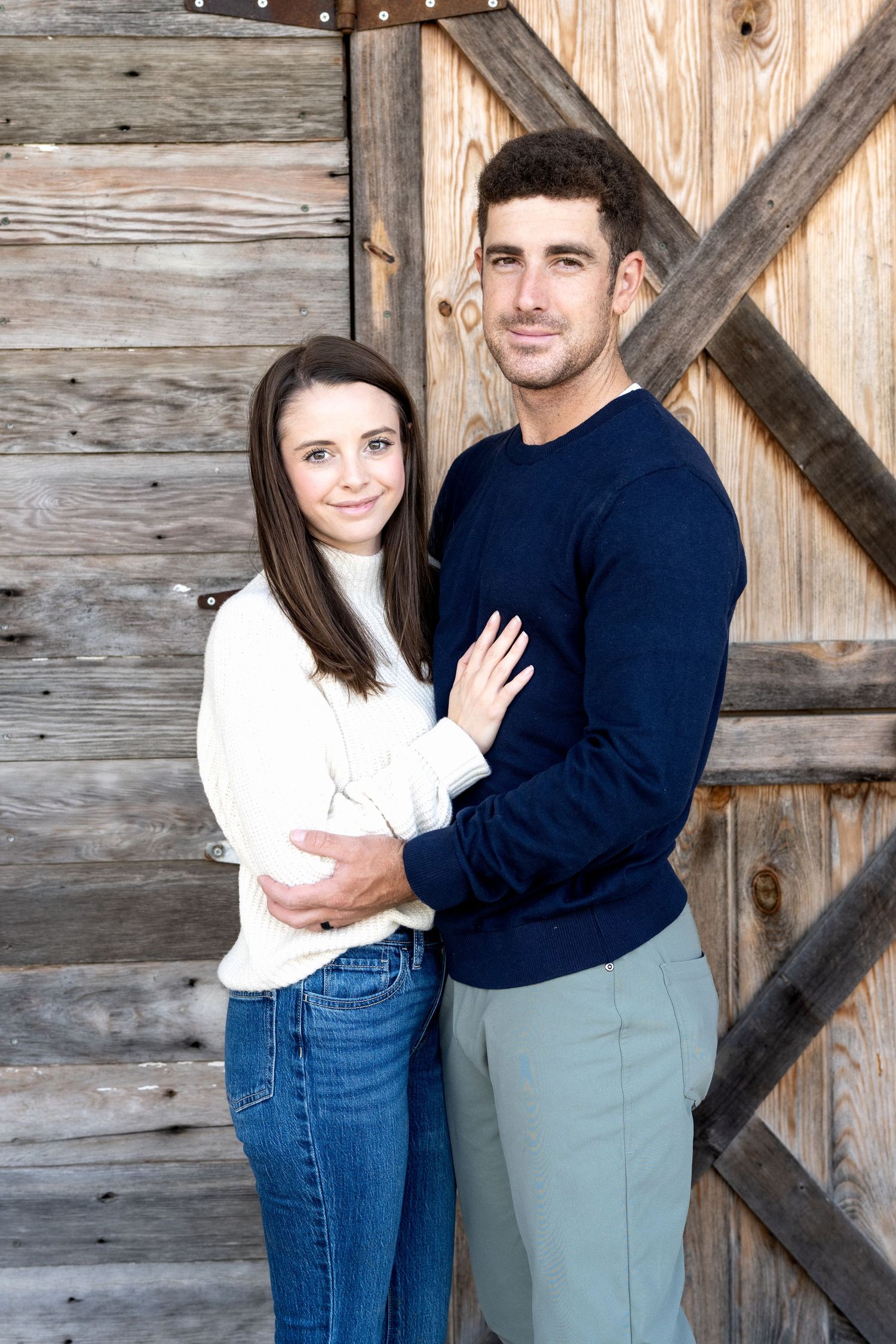 A young couple embracing in front of a rustic wooden barn door, wearing casual sweaters and jeans.