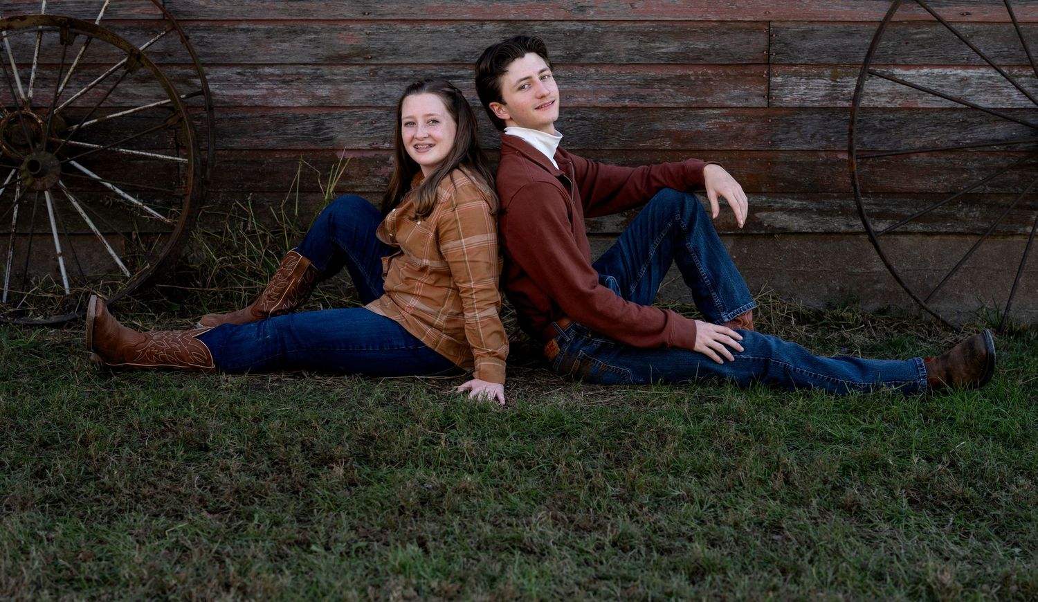Two smiling teenagers sit back to back on green grass in front of a rustic barn wall and old wagon wheels.