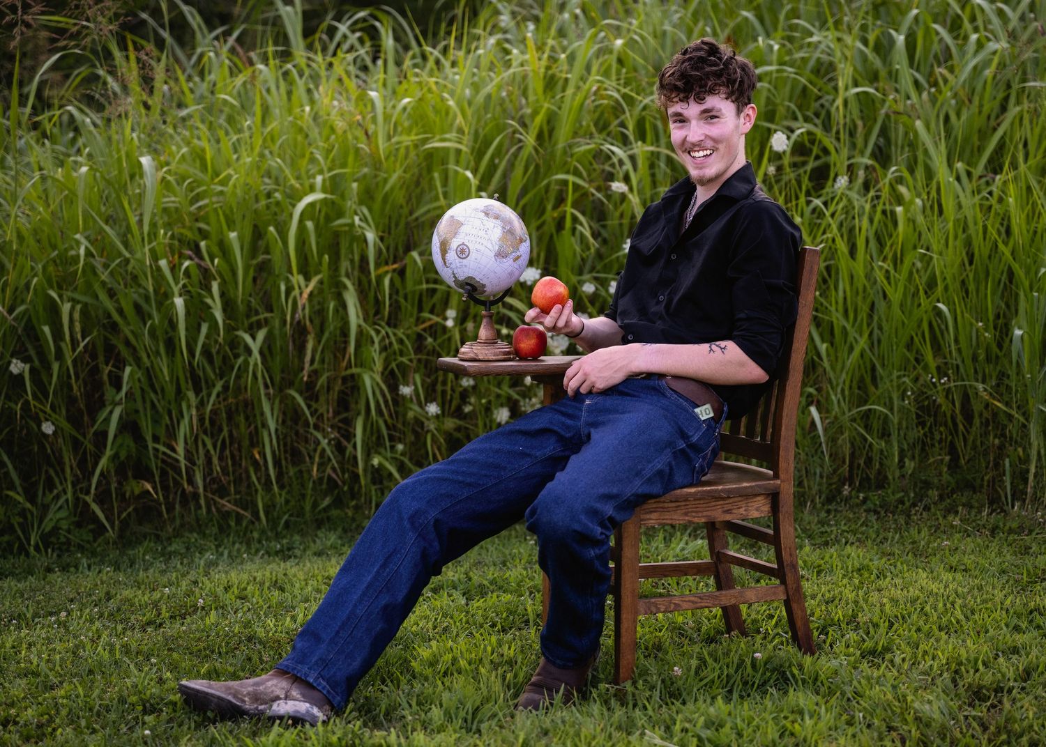 portrait of a  teen boy sitting in wooden chair outdoors with an apple and globe on small table.