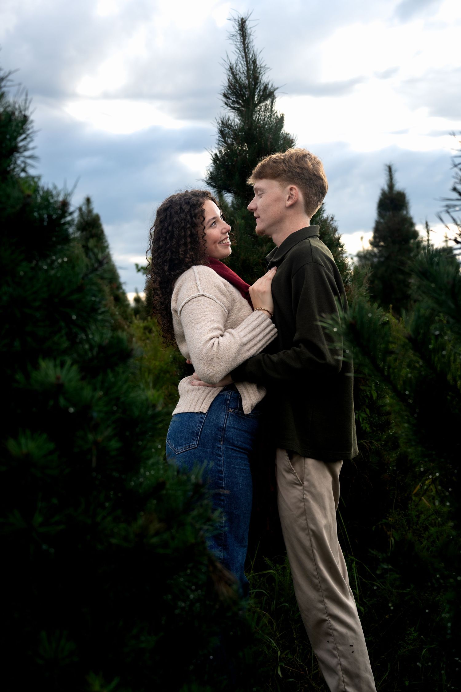 A couple embraces romantically among lush green Christmas trees under a cloudy sky.