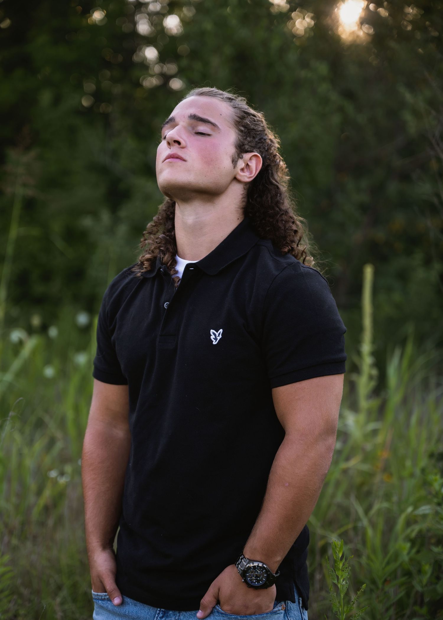portrait of a young man with long curly hair poses eyes closed in outdoor setting.