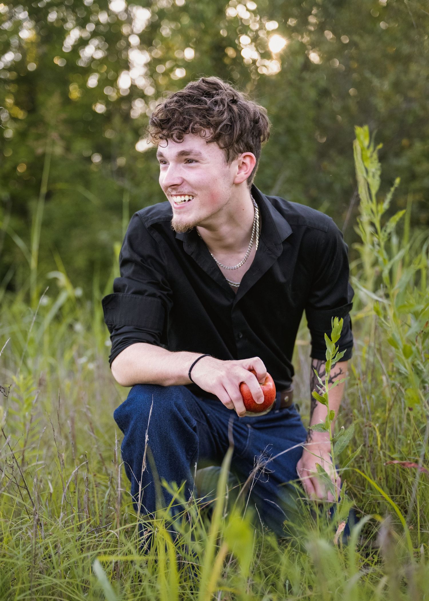 portrait of a Smiling young man with arm tattoo crouching in tall green grass holding a red apple.