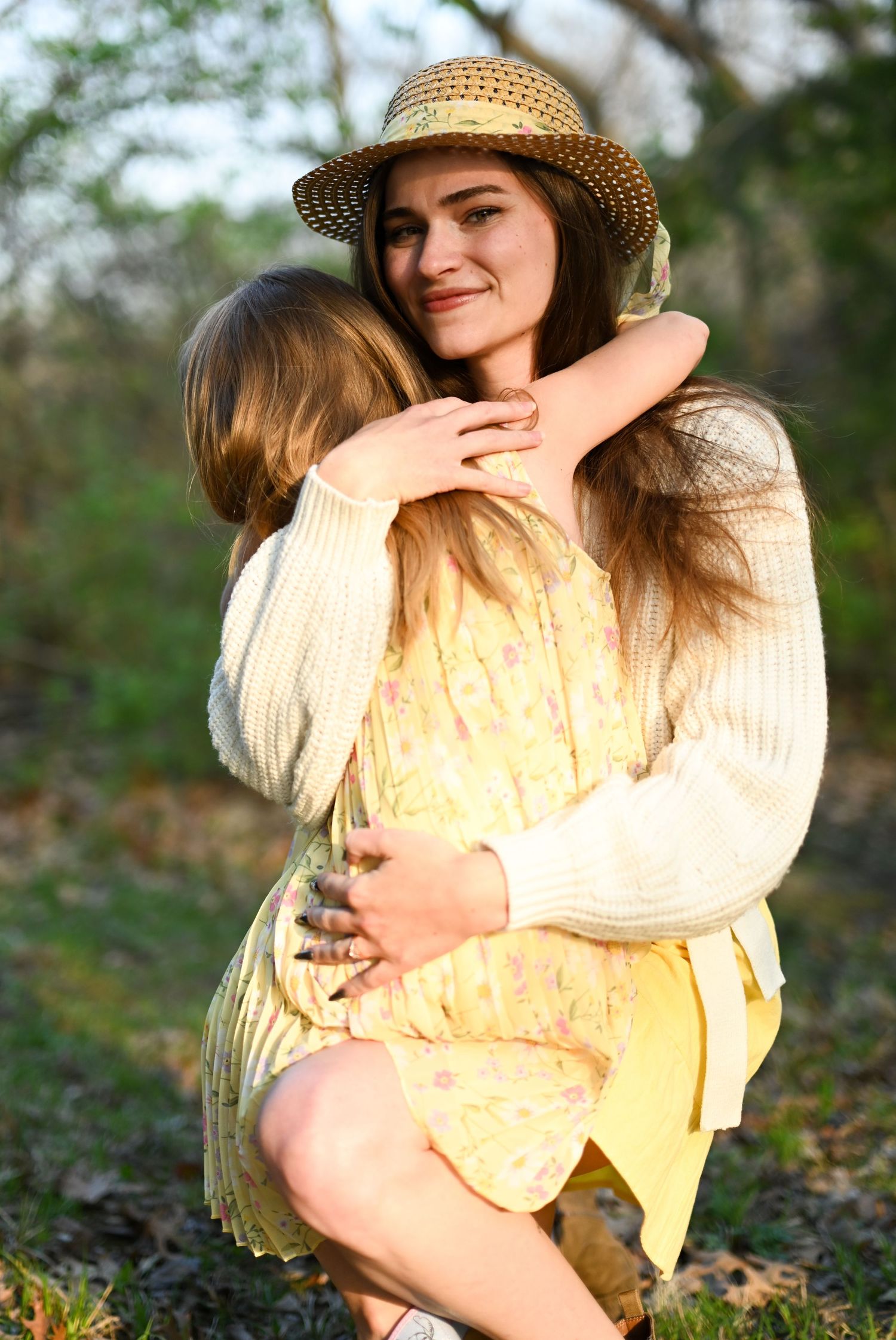 Young woman in straw hat hugging a little girl outdoors in golden sunlight.