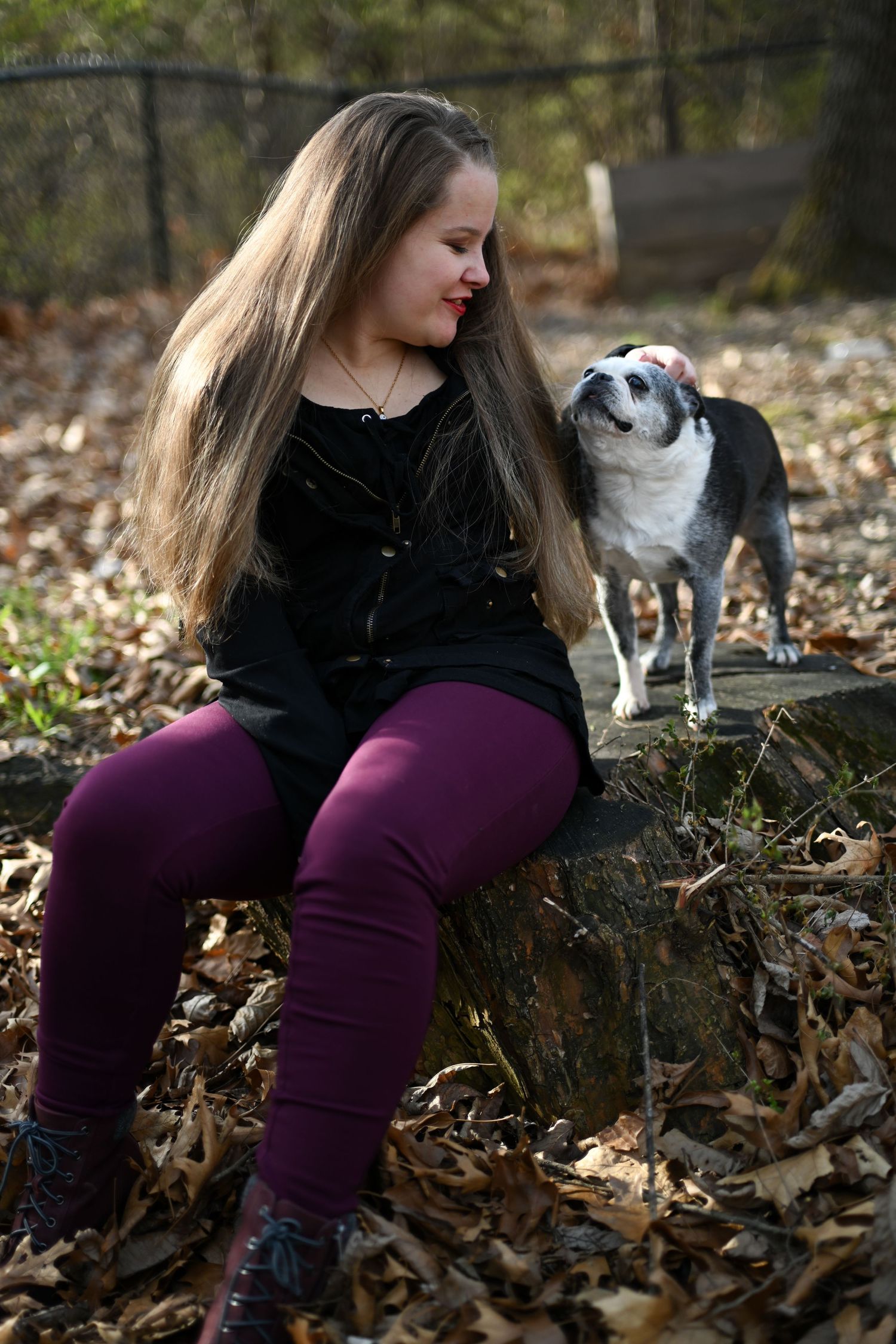 Young woman in black outfit sitting on stone wall with Boston Terrier dog outdoors.
