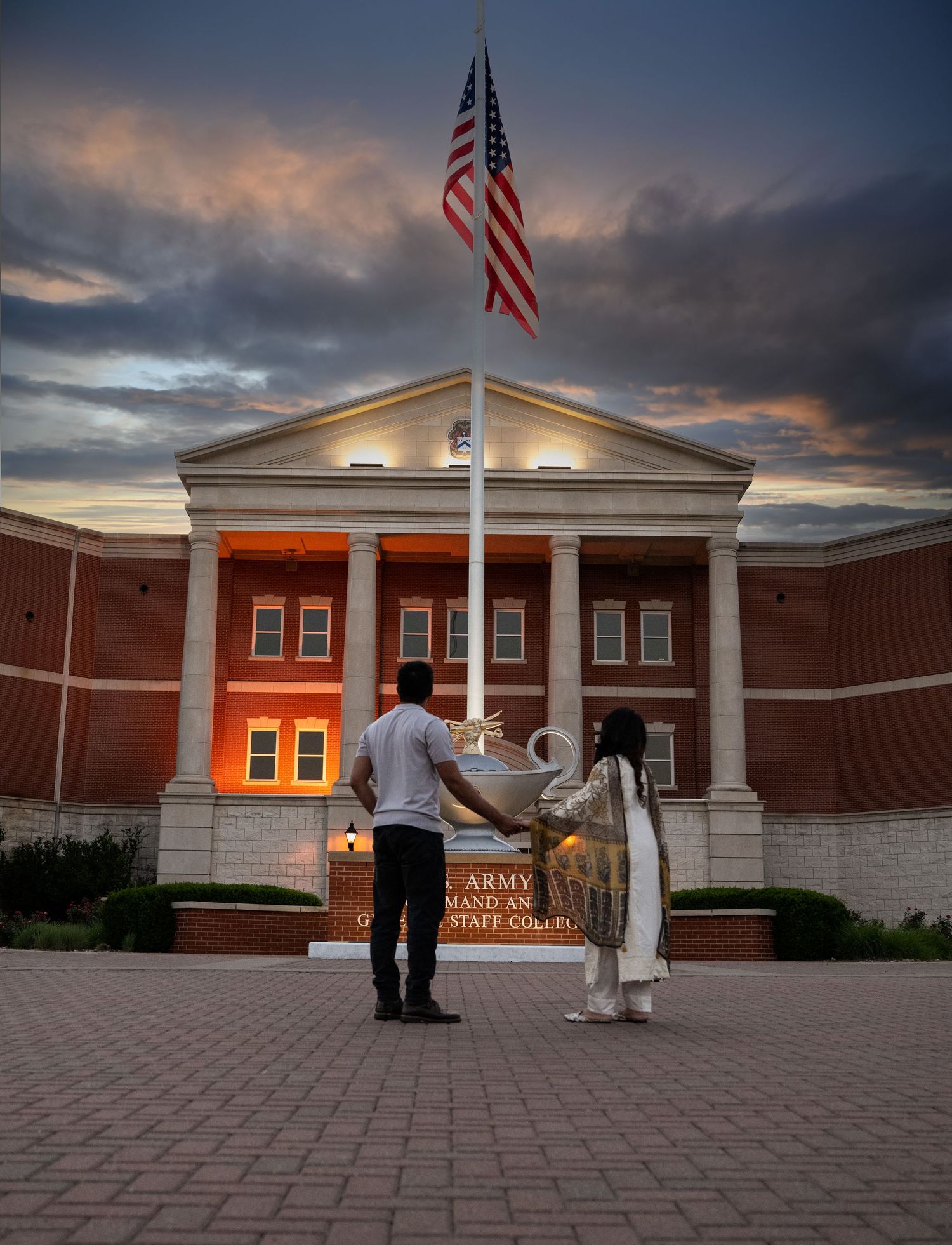 A couple stands before a US Army Command and Staff College building at dusk under an American flag.