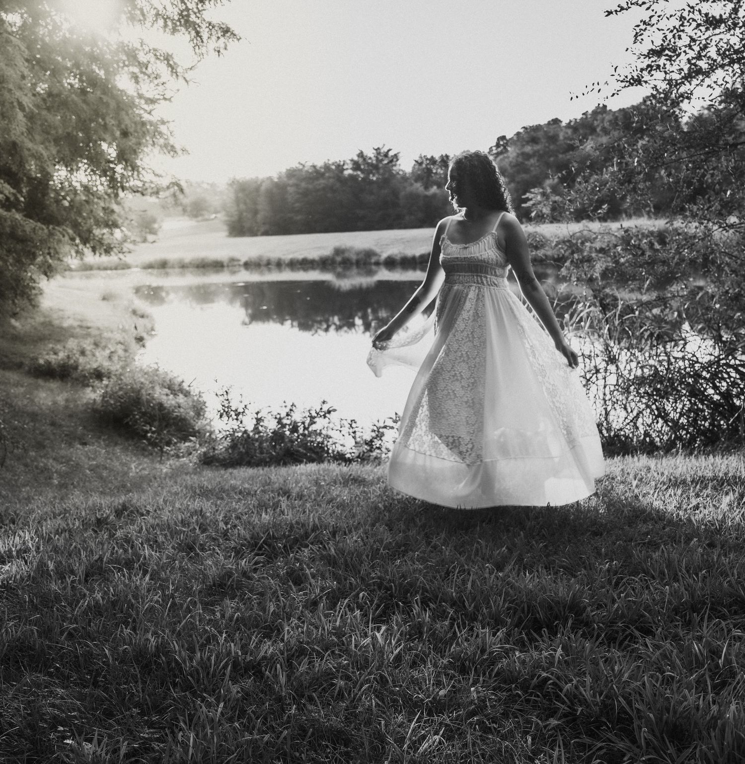 lifestyle portrait of A woman in a white gown standing by a tranquil pond surrounded by lush trees in a dreamy black and white photo.