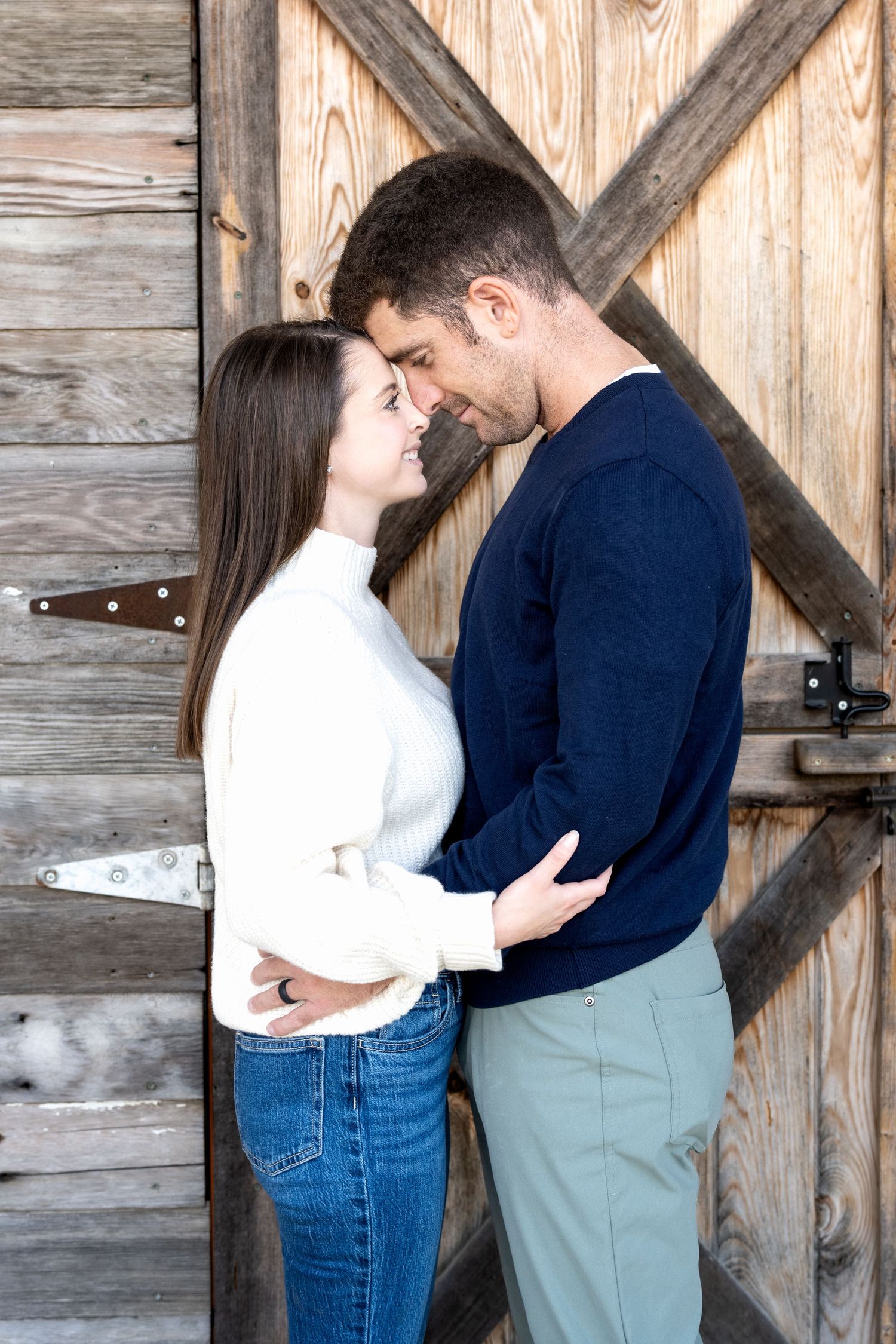 Couple embracing romantically in front of rustic wooden barn doors, woman in white sweater and jeans, man in navy top.