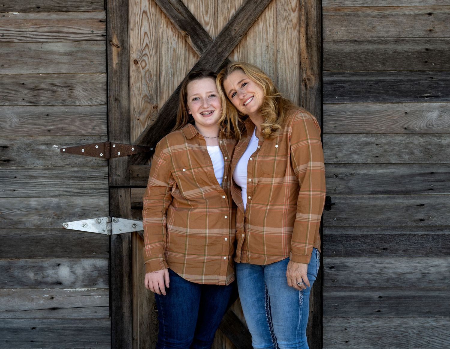 Two smiling women in matching orange plaid flannel shirts and jeans pose side by side in front of a rustic weathered wooden barn door.