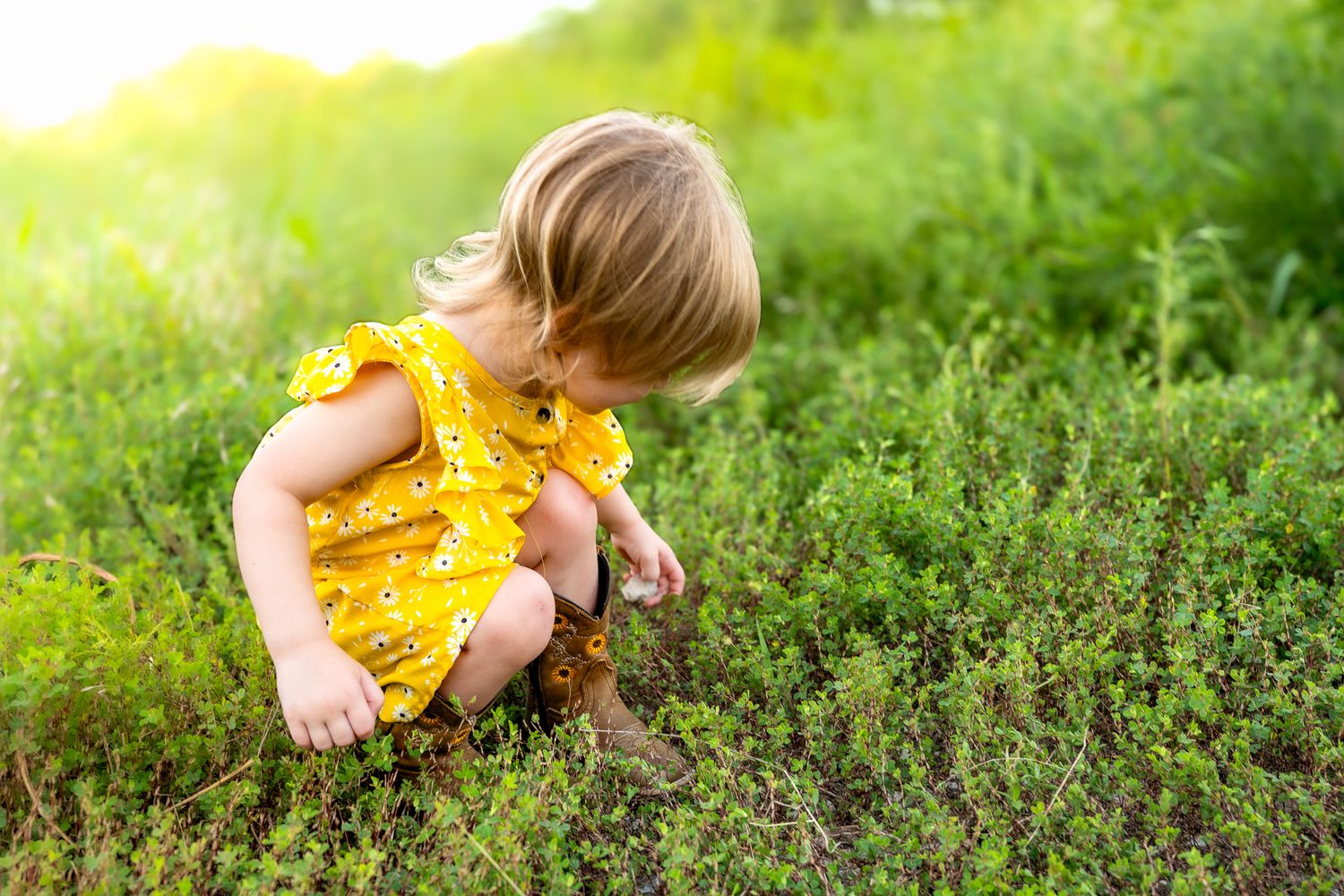 Toddler girl in yellow floral romper exploring green bushes outdoors in sunny garden setting. faceless family photography in Leavenworth, KS