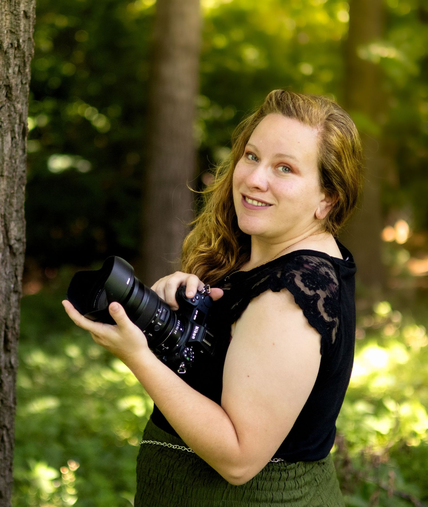 Woman photographer smiling and holding a camera outdoors in a lush green forest setting.