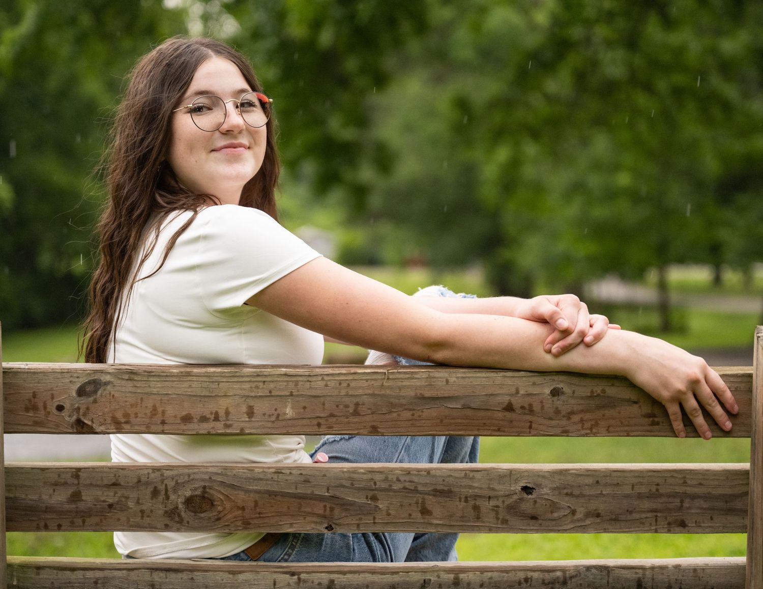 portrait of a Smiling teenage girl with glasses resting arms on wooden park bench surrounded by greenery.