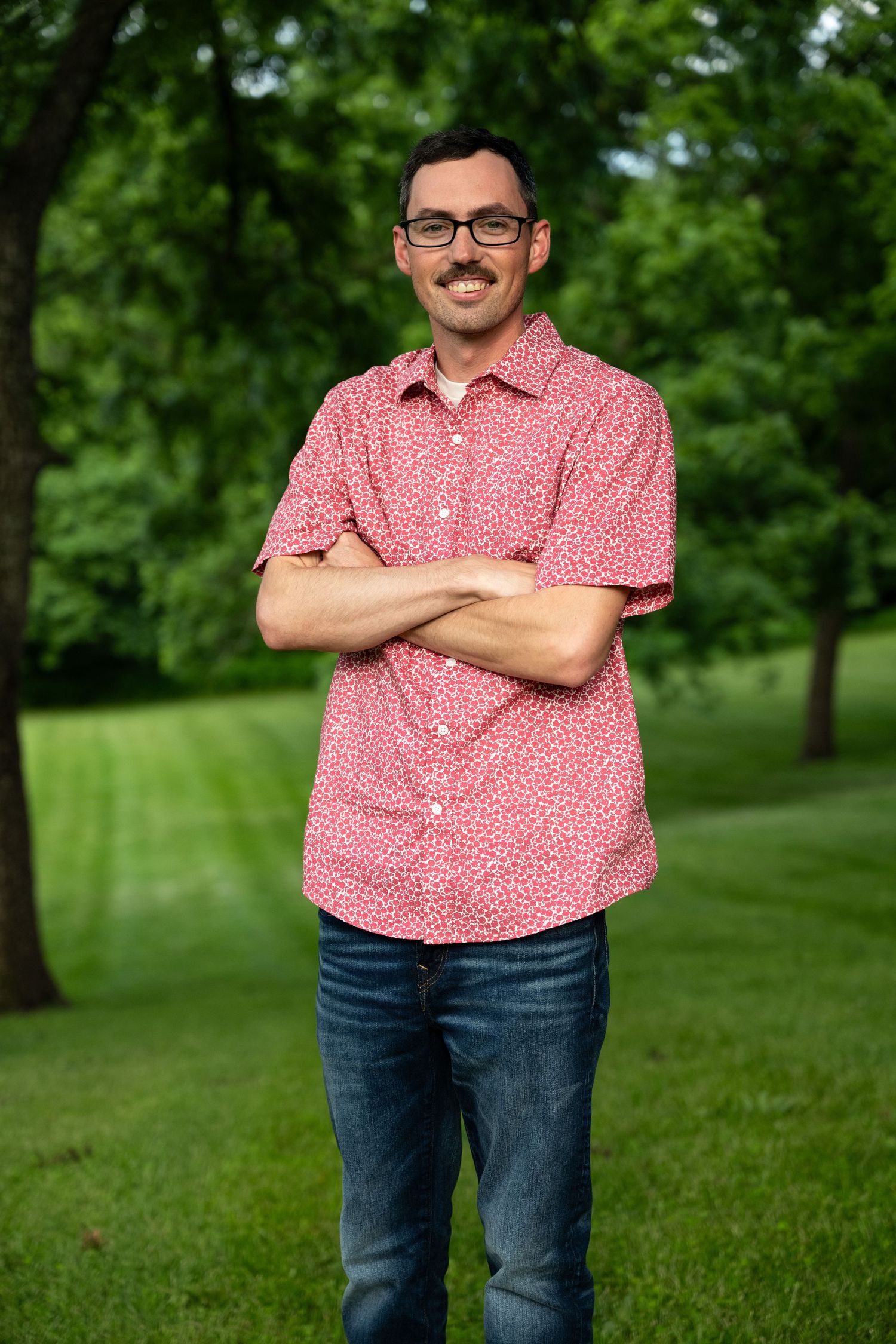 A smiling man with glasses and a mustache stands with arms crossed in a green park.