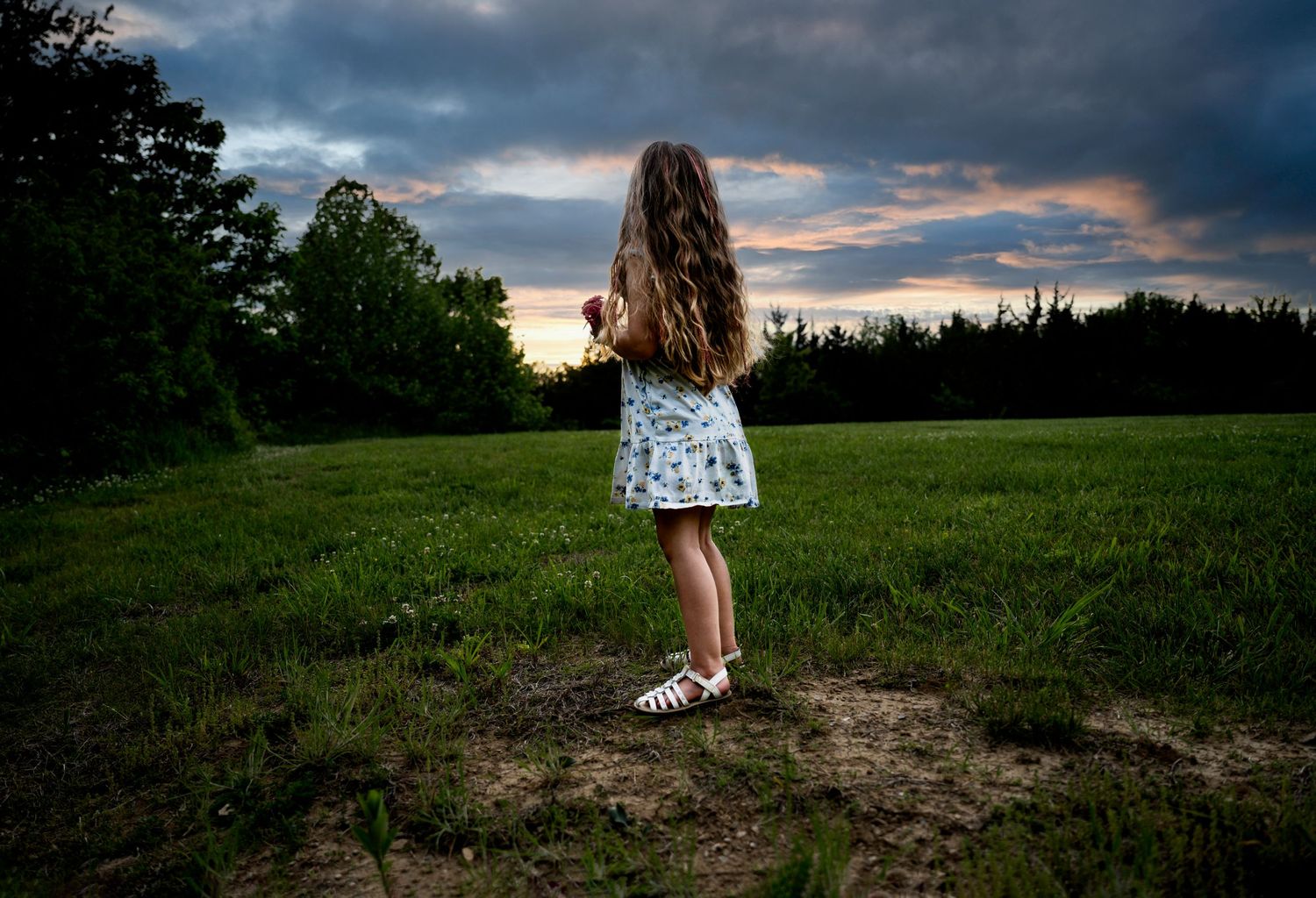 A young girl with long curly hair stands in a field at sunset wearing a light summer dress.