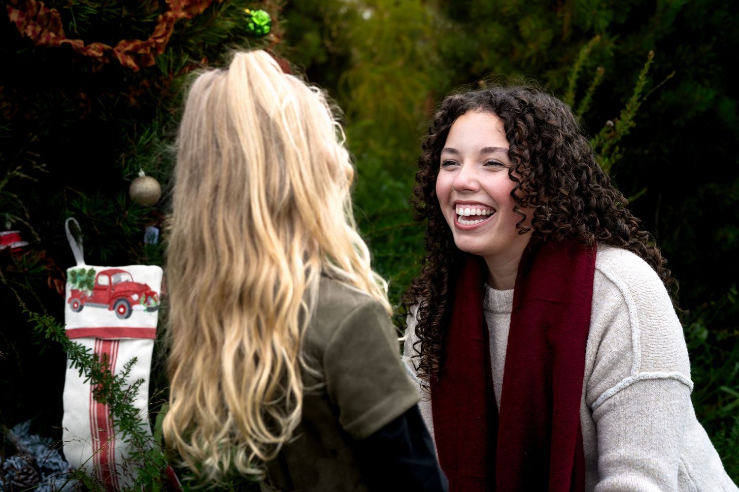 Two women laughing together outdoors near a decorated Christmas tree with stockings and ornaments.