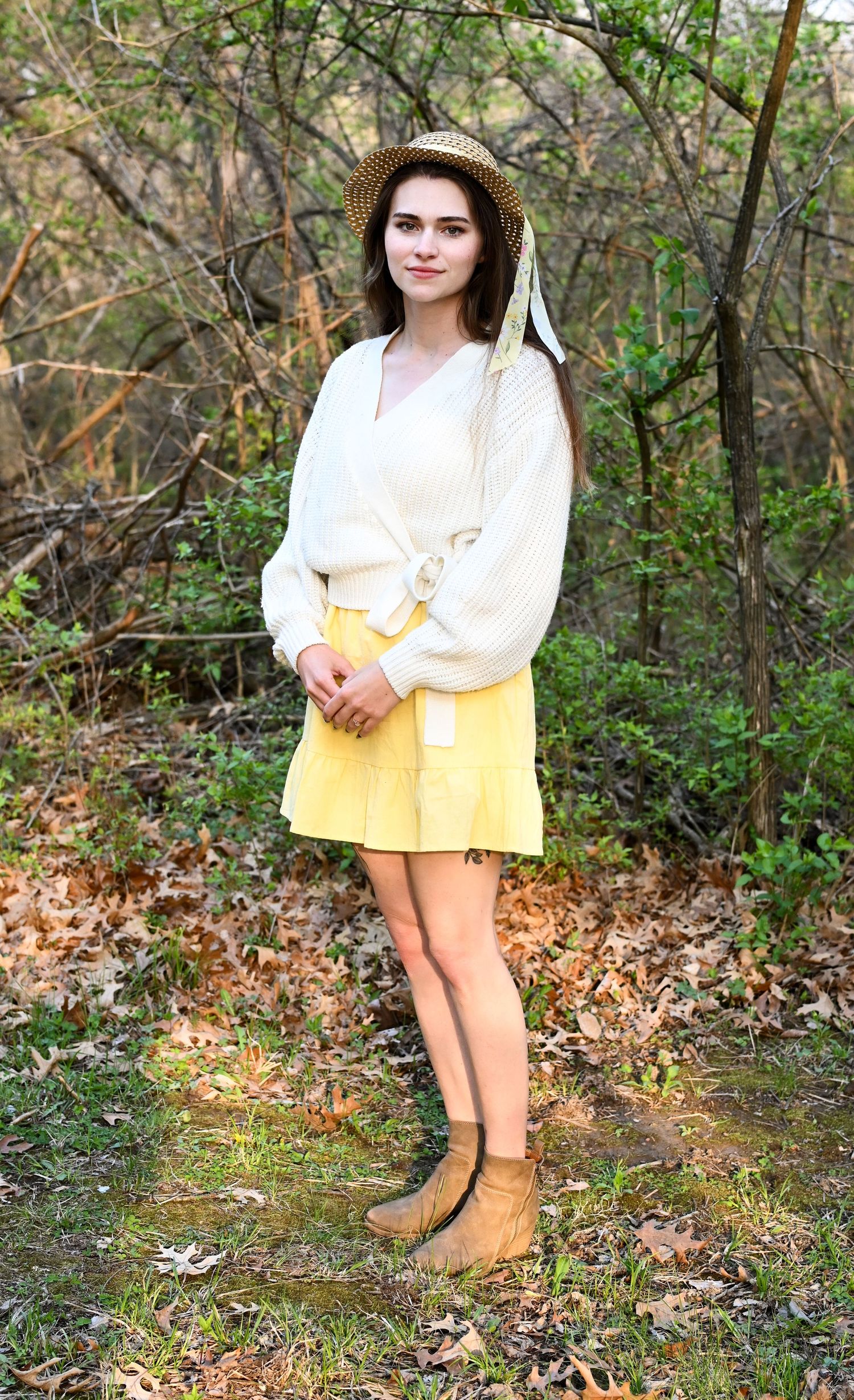 Young woman in white sweater, yellow skirt, and straw hat posing in autumn woods.
