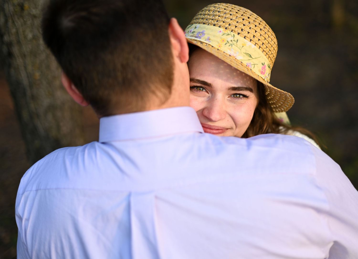 A smiling young woman in a floral straw hat embraces a man in a white shirt outdoors.