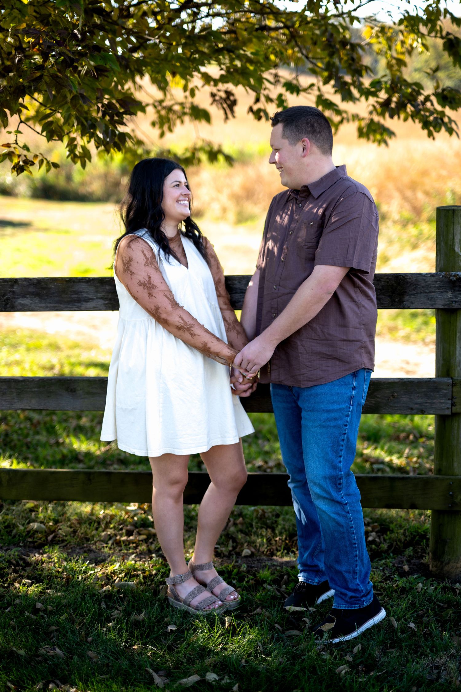 A couple holds hands and smiles at each other by a wooden fence in a sunny outdoor setting.