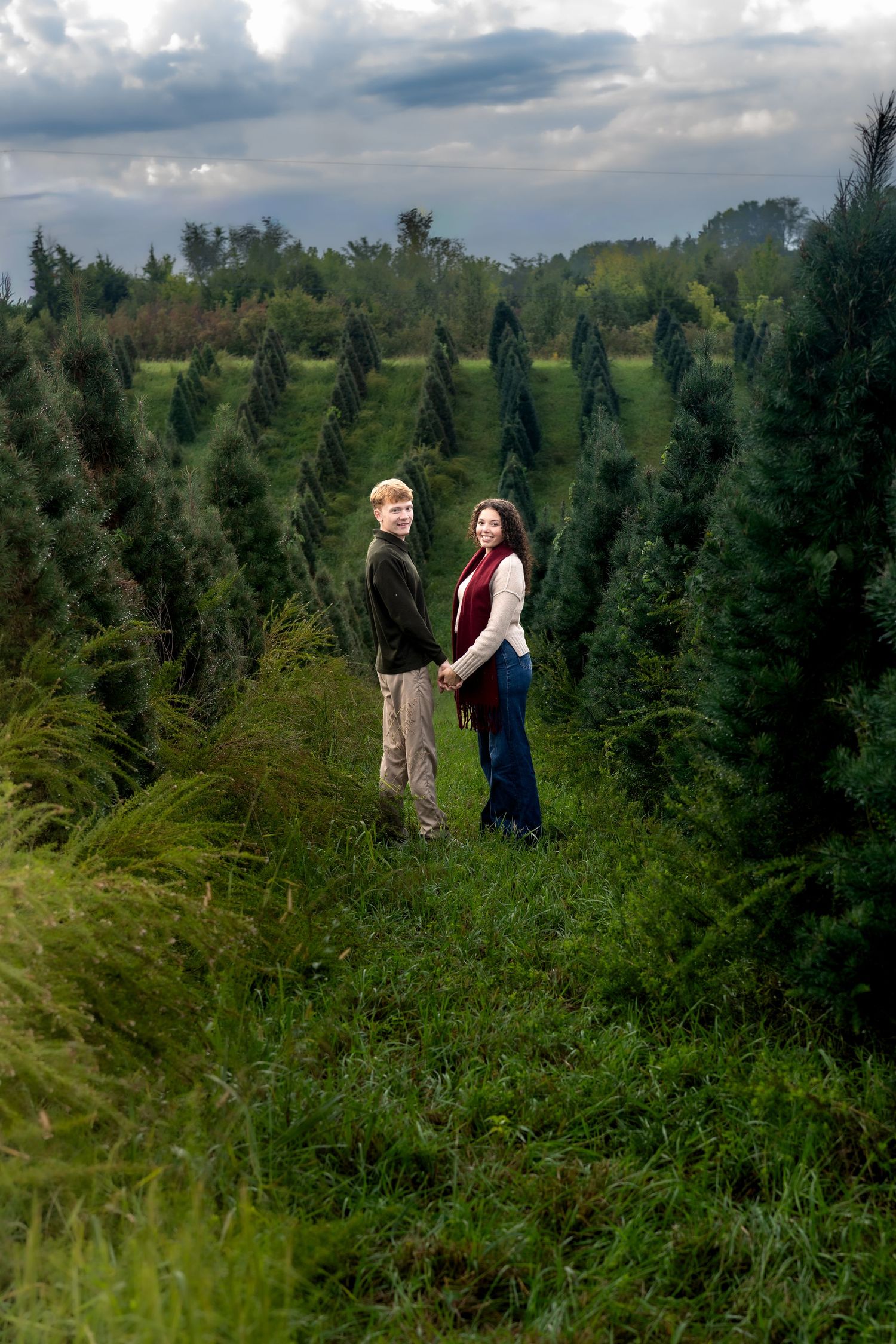 Couple holding hands among rows of green Christmas trees at a tree farm under cloudy skies.