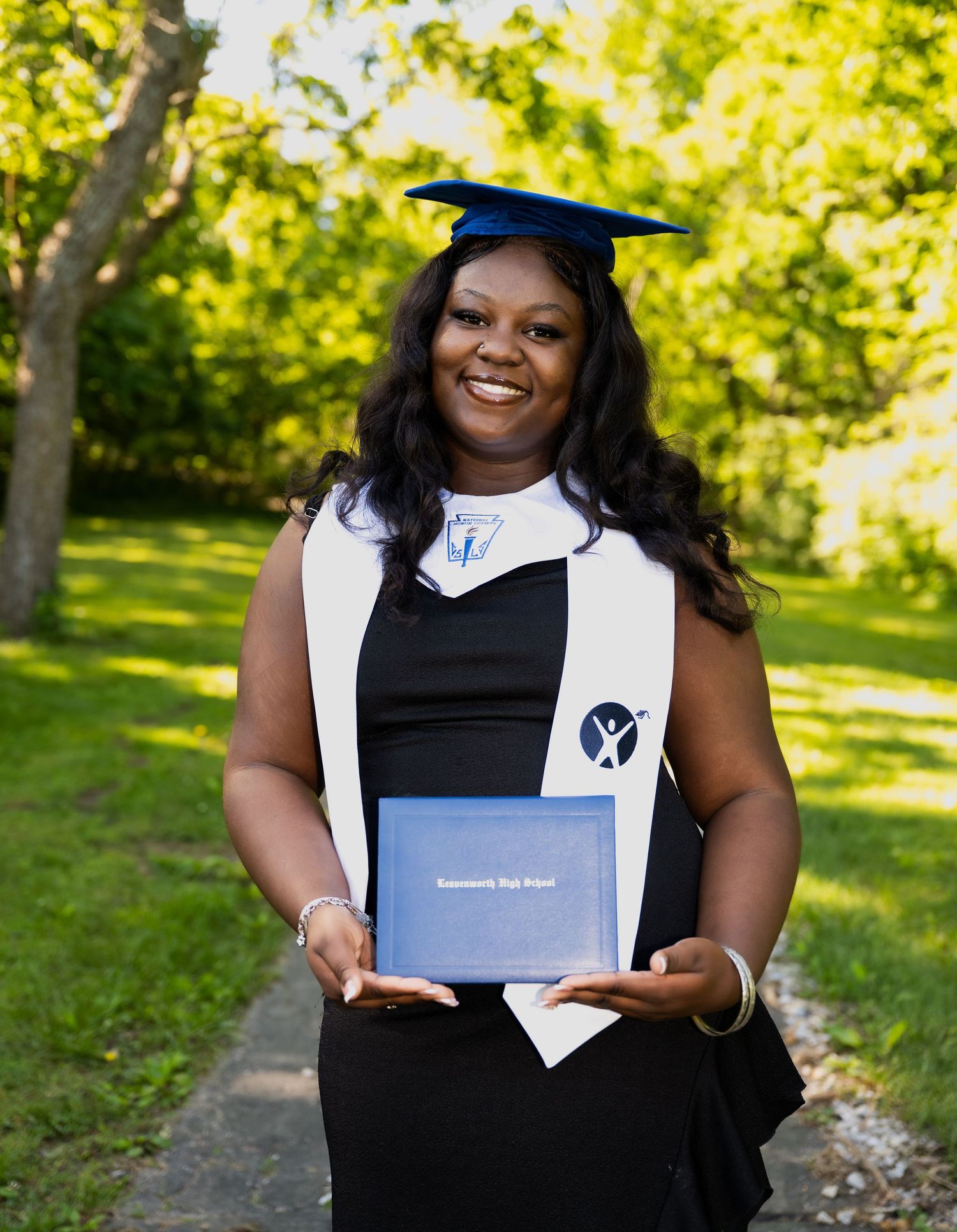 portrait of a Proud graduate in blue cap and gown holding diploma standing on a sunny tree-lined outdoor path.