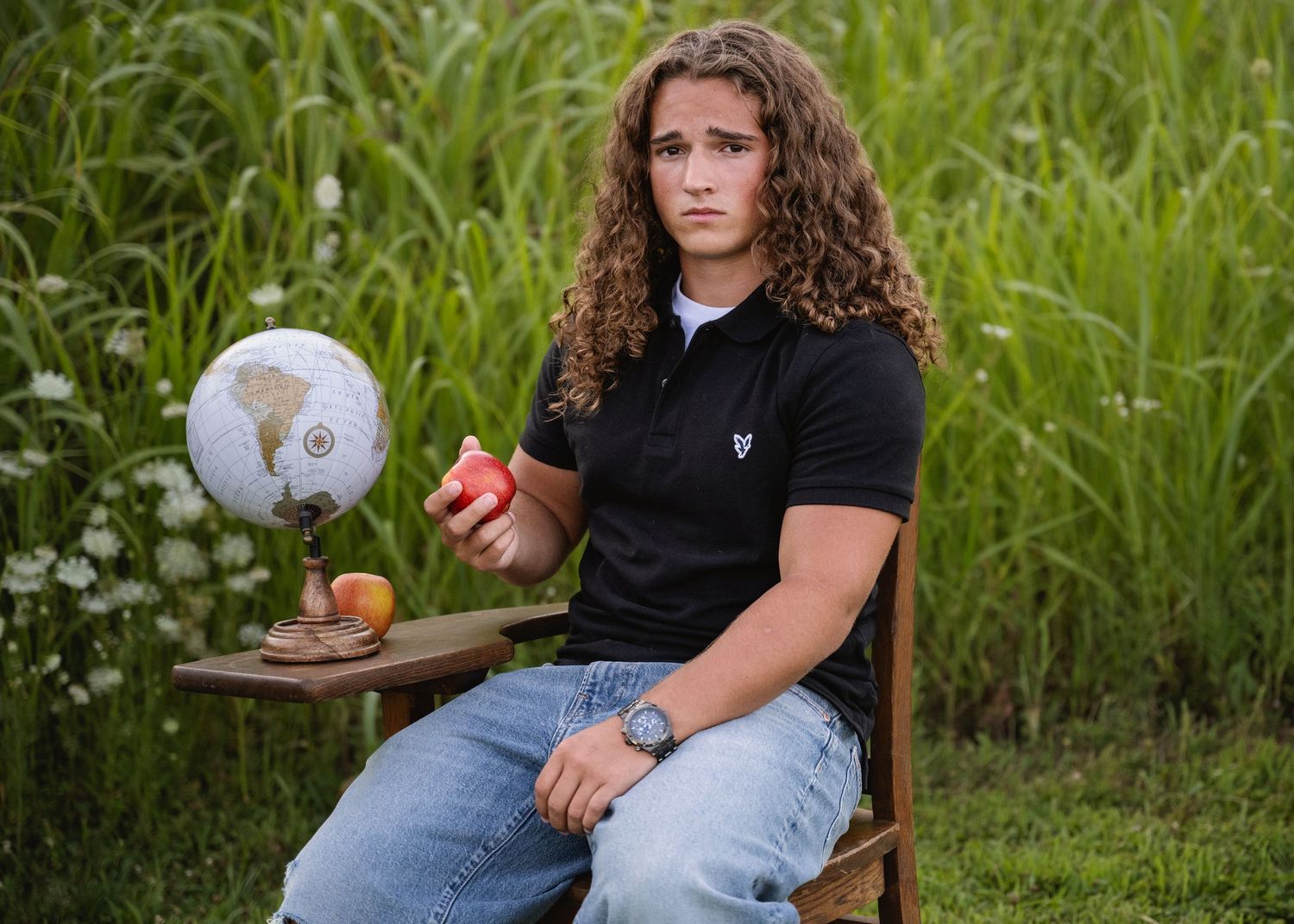 portrait of a Teen with curly hair sitting in wooden chair outdoors holding apple with globe on table.