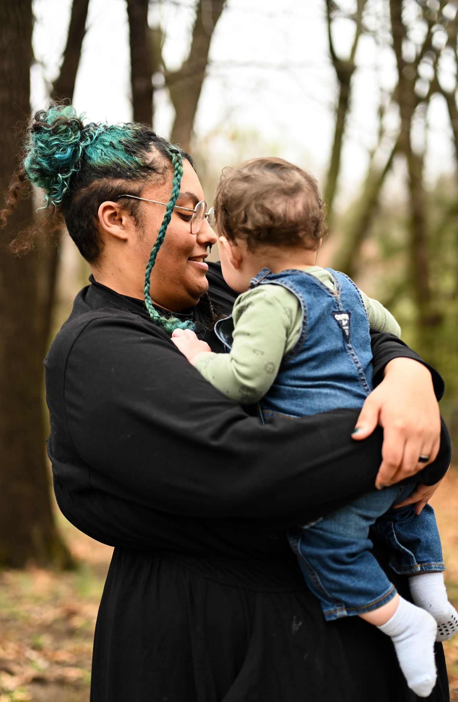 Loving mother with colorful braids holding baby in denim overalls nose-to-nose in wooded park.