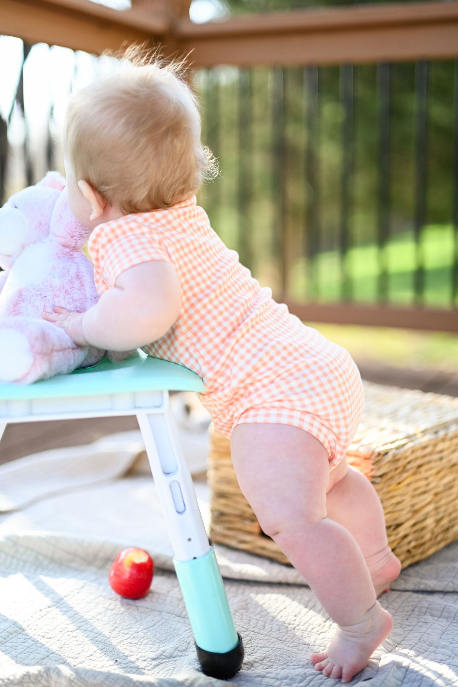 Baby in pink gingham romper standing on a wicker basket on a sunny patio.