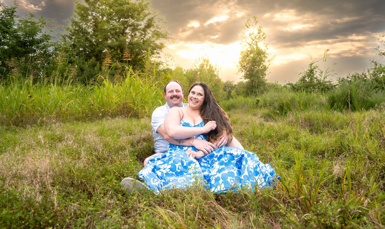 lifestyle portrait of a Couple sitting together in a lush green meadow at sunset, woman in blue floral dress, man in white shirt.