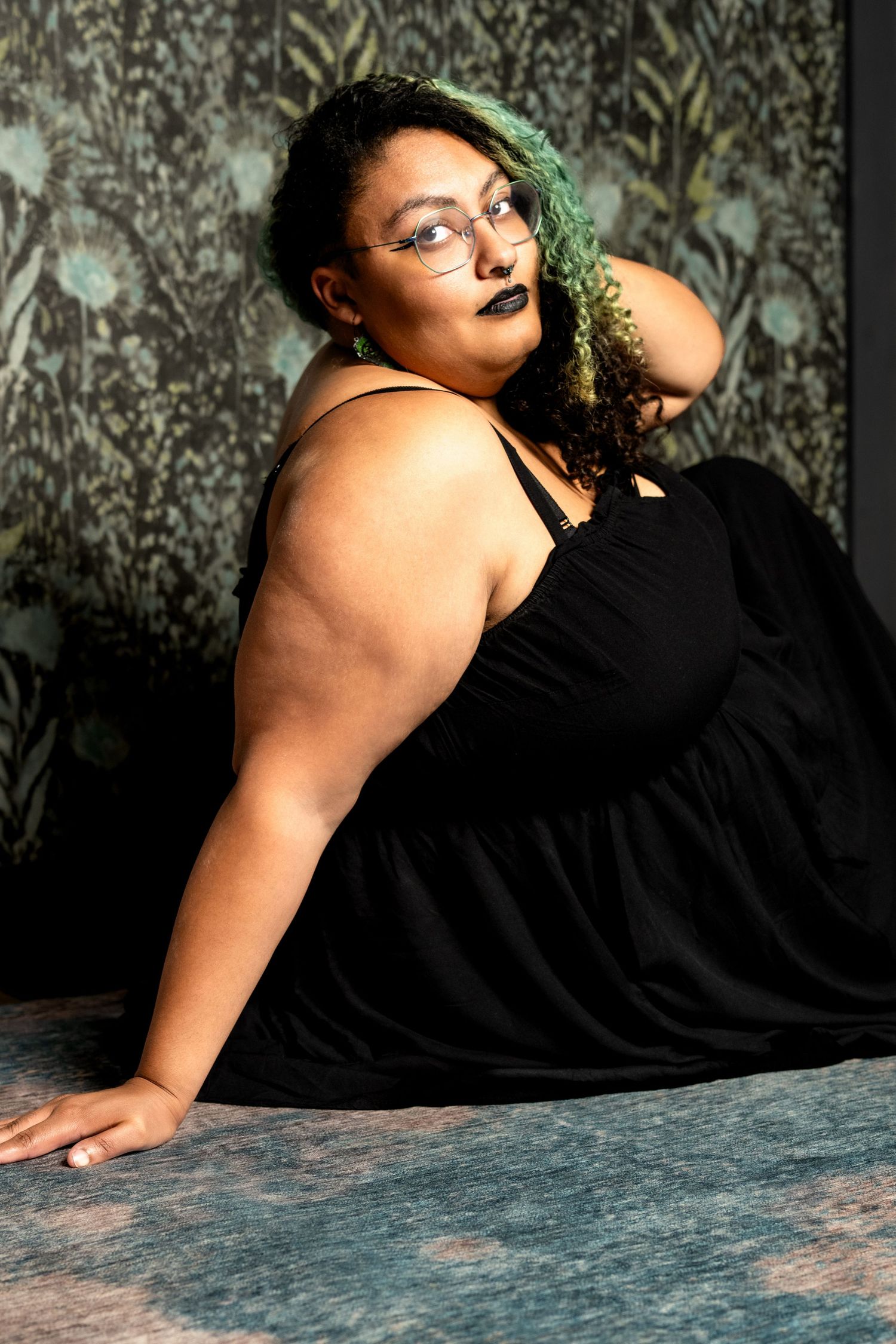 Plus-size woman in black dress with green hair and glasses poses confidently against floral wallpaper.